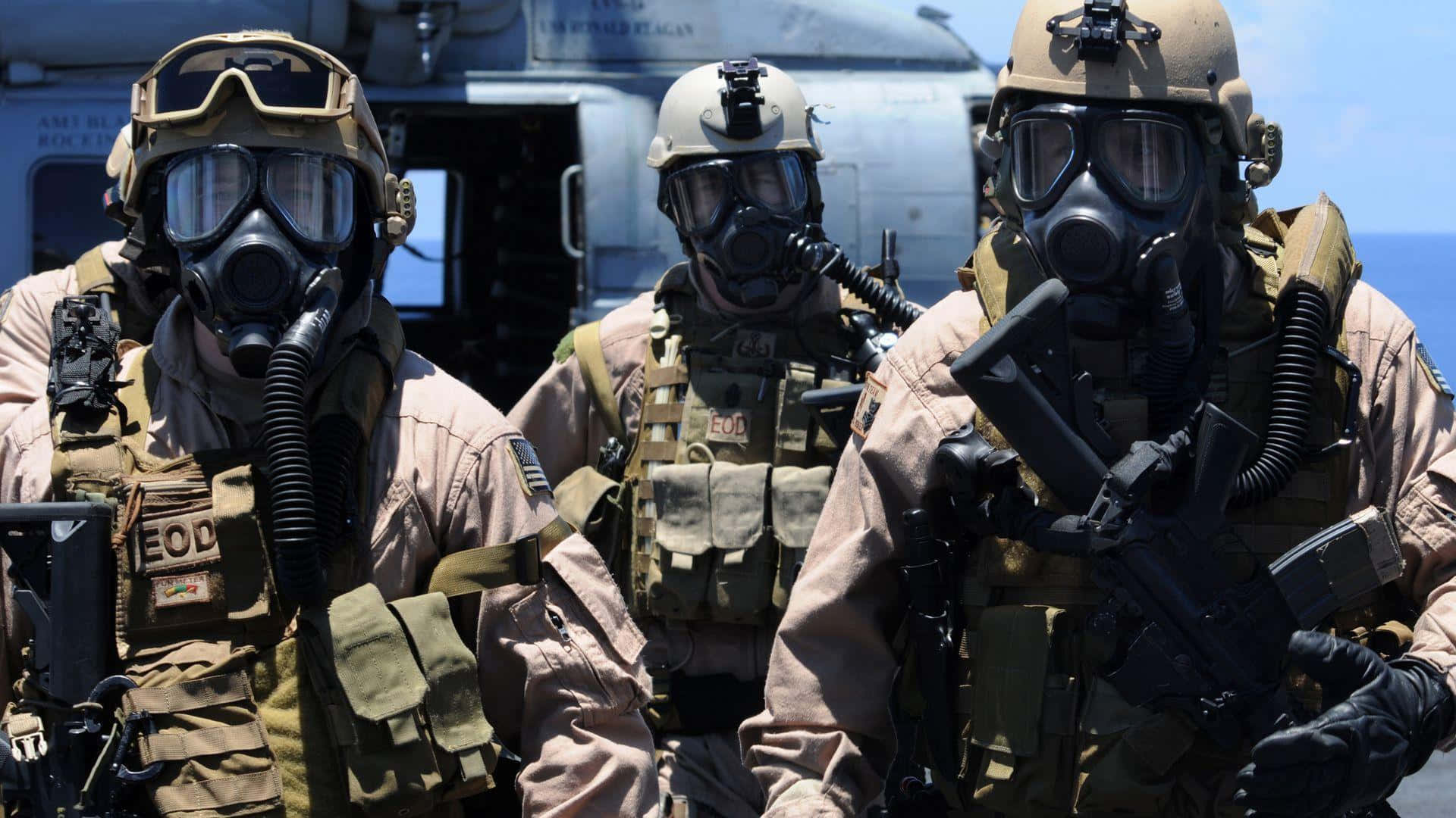 Special Forces 桌布 壁紙 — 三名身穿全套戰術裝備和防毒面具的EOD士兵在明亮的日光下警覺地站在軍用直升機附近。