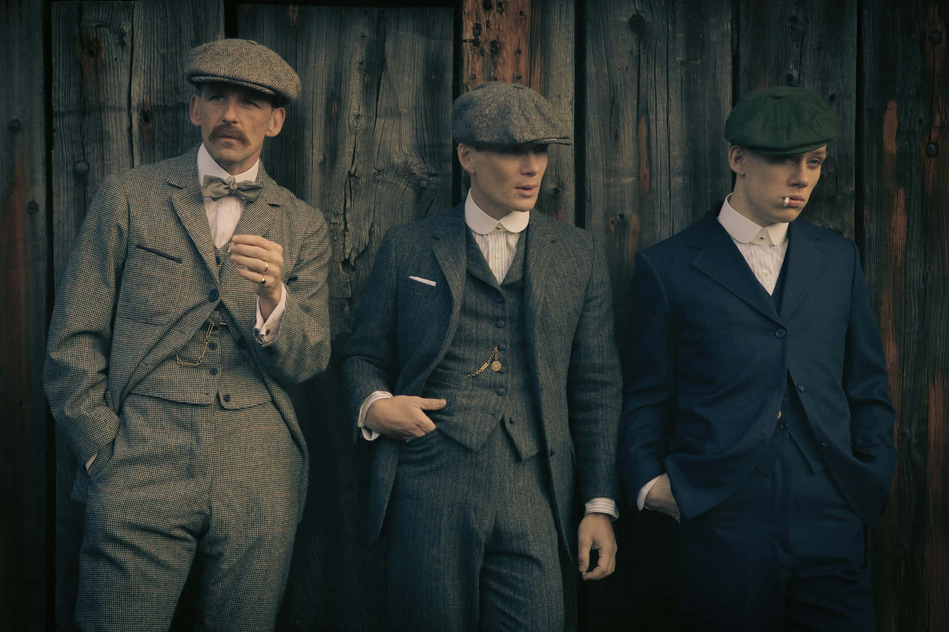 三名身穿復古西裝和扁帽的男子靠在木牆上。 — Peaky Blinders 壁紙