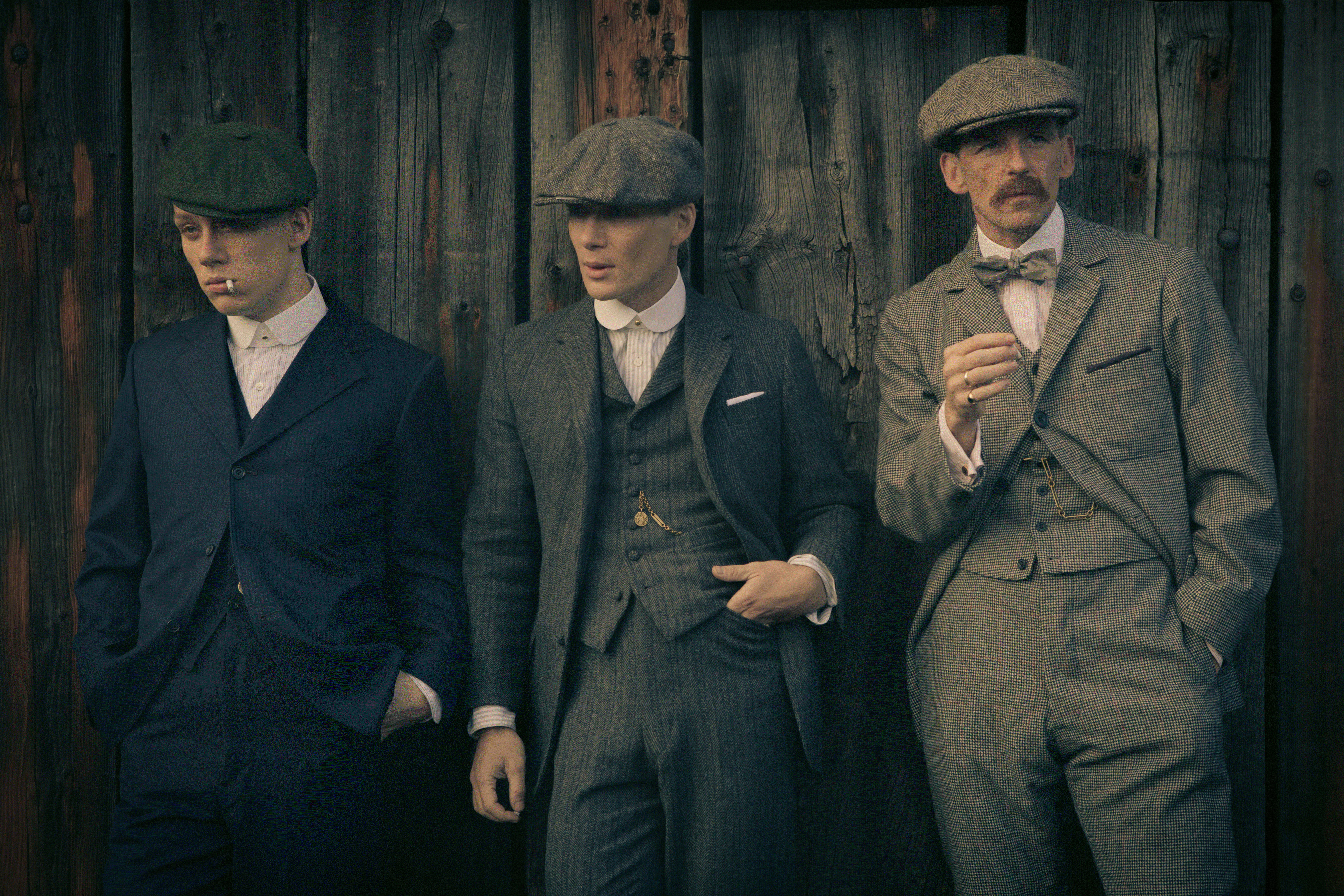 三名身穿復古西裝和扁帽的男子靠著木牆站立。 — Peaky Blinders 壁紙
