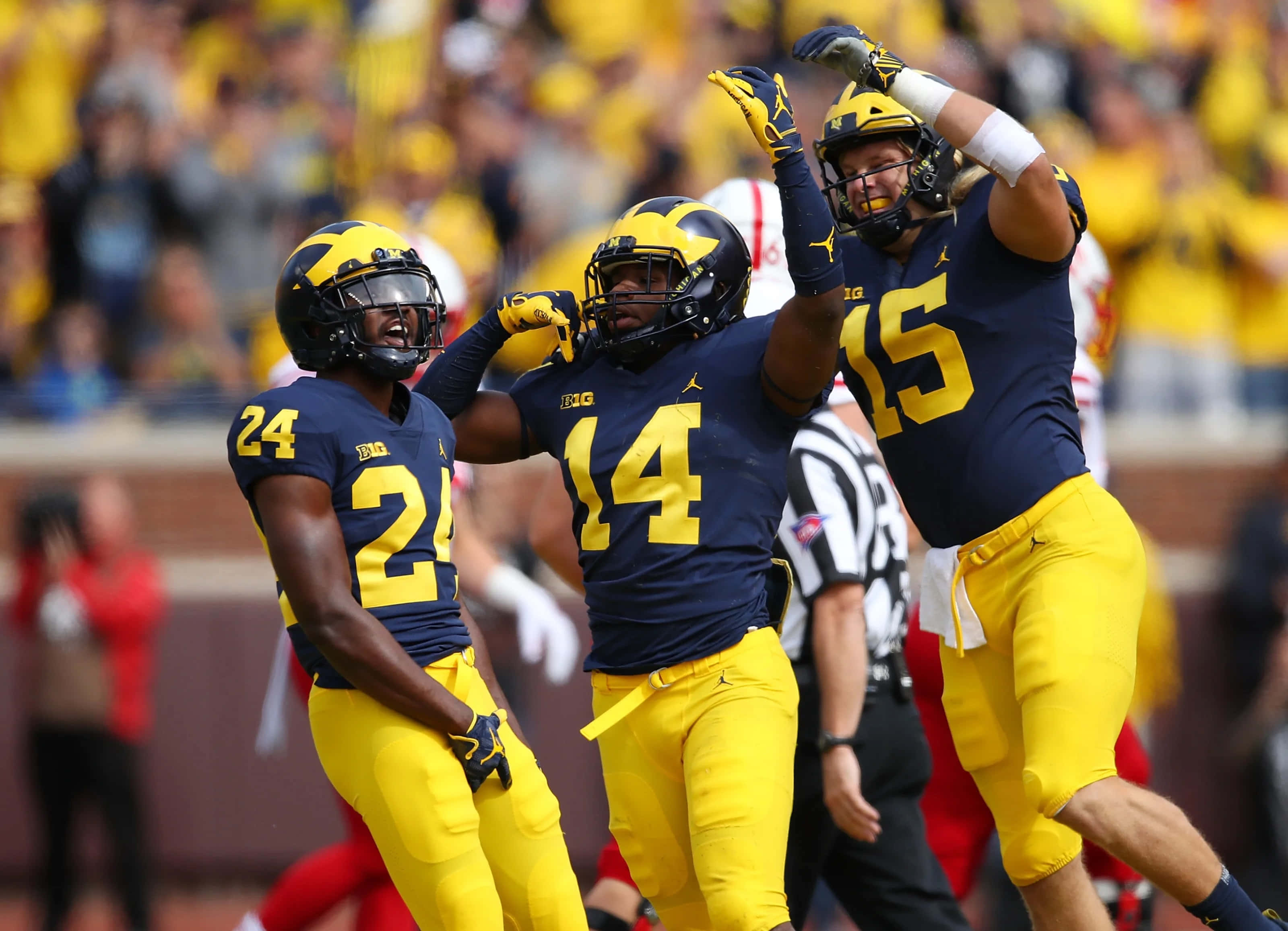 Michigan Football 桌布 壁紙 — 三名身穿海軍藍和黃色球衣的密西根大學美式足球員在陽光明媚的比賽日以擊掌和舉臂慶祝一次進攻。