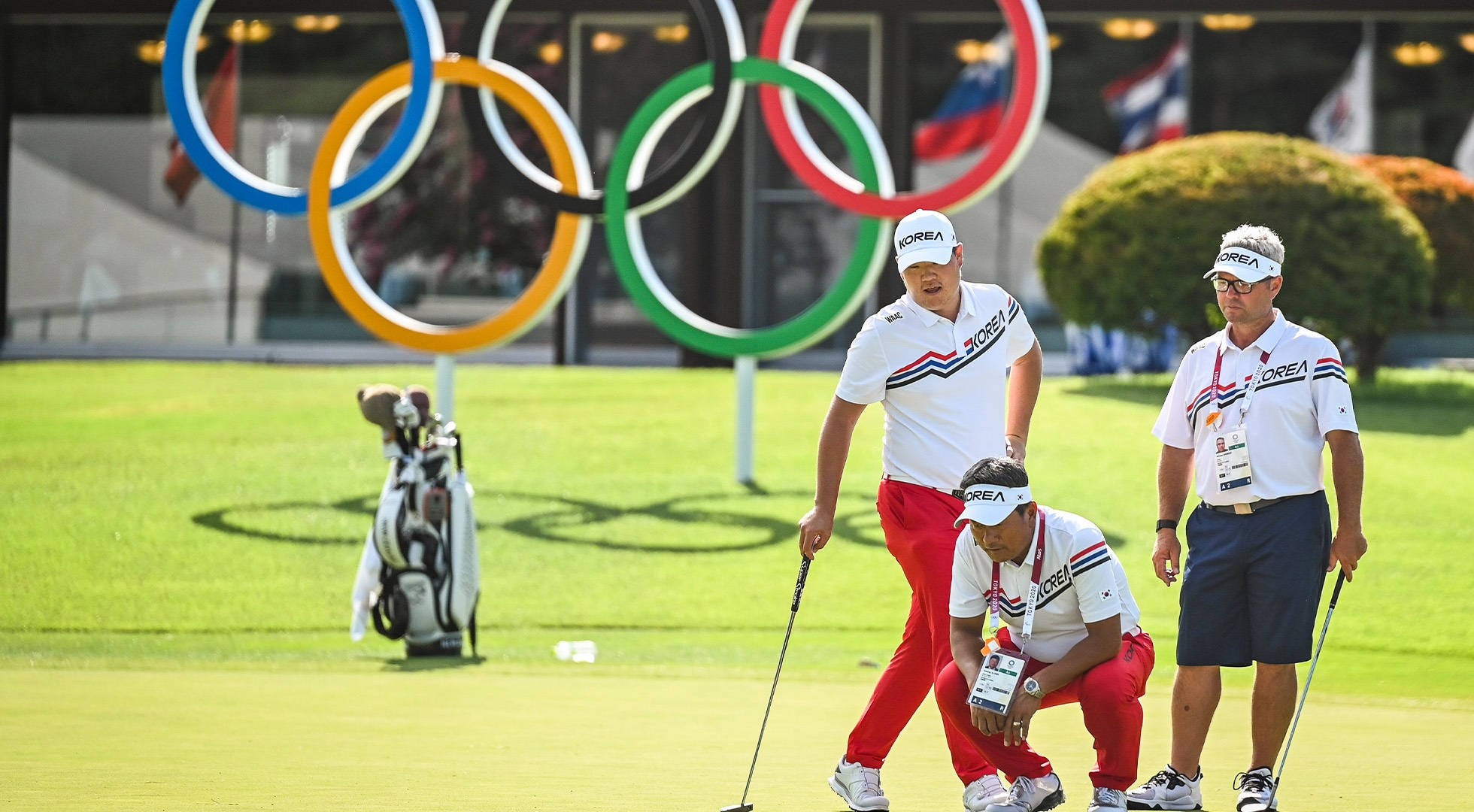 Olympics 桌布 壁紙 — 三名身穿白色和紅色制服的高爾夫球手在陽光普照的果嶺上分析推桿，背後聳立著奧運五環。