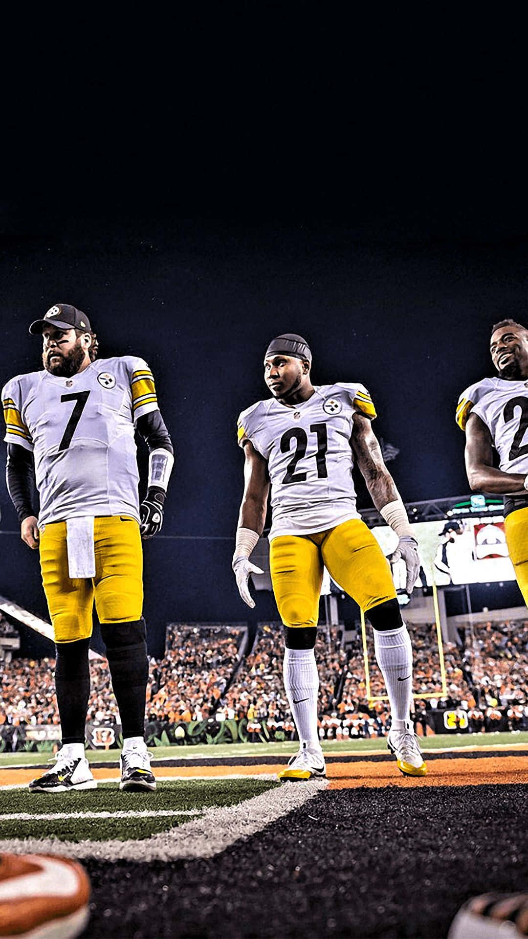 Steelers iPhone 桌布 壁紙 — 三名身穿白色和黃色球衣的匹茲堡鋼人隊美式足球員在夜間球場燈光下站在場上，背景有觀眾。