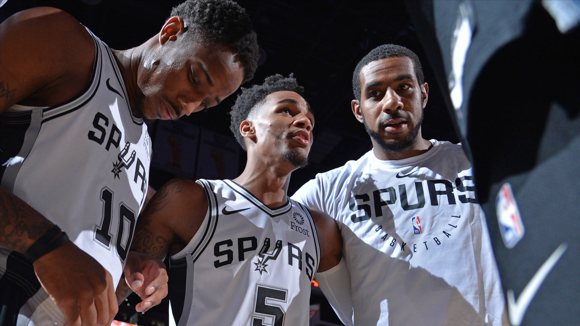 Dejounte Murray 桌布 壁紙 — 三名身穿白色球衣的聖安東尼奧馬刺籃球運動員在明亮的場館燈光下於比賽中緊密圍攏。