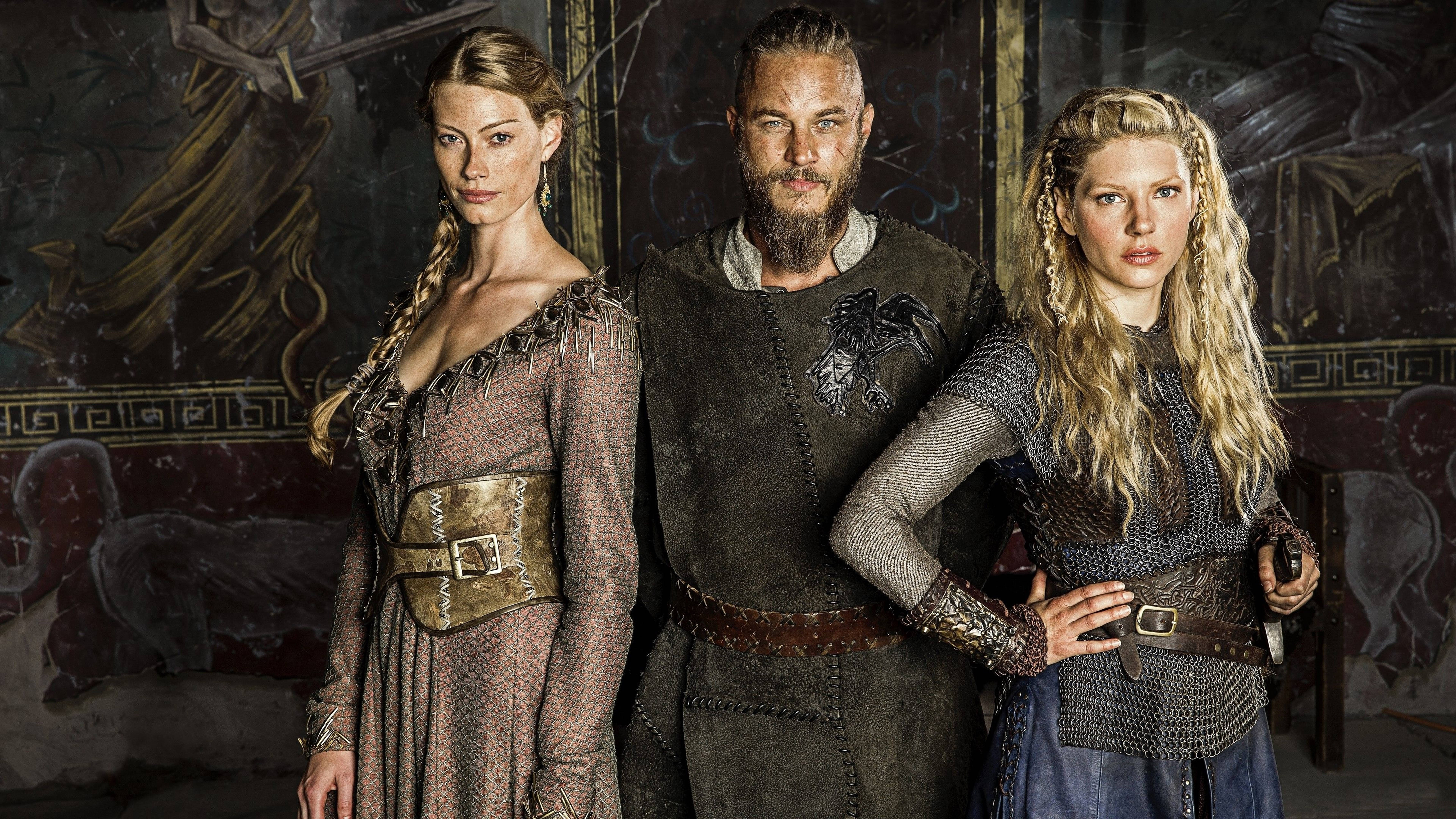 Vikings 桌布 壁紙 — 三名身穿精細盔甲的維京時代戰士，莊嚴地站在一幅裝飾著壁畫的石牆前。