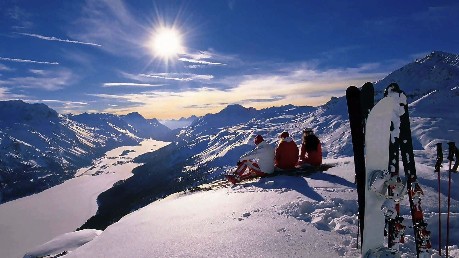 Winter Sports 桌布 壁紙 — 三名身穿紅色外套的滑雪者坐在白雪覆蓋的山脊頂端，沐浴在明亮的陽光下，俯瞰廣闊的冰凍山谷。