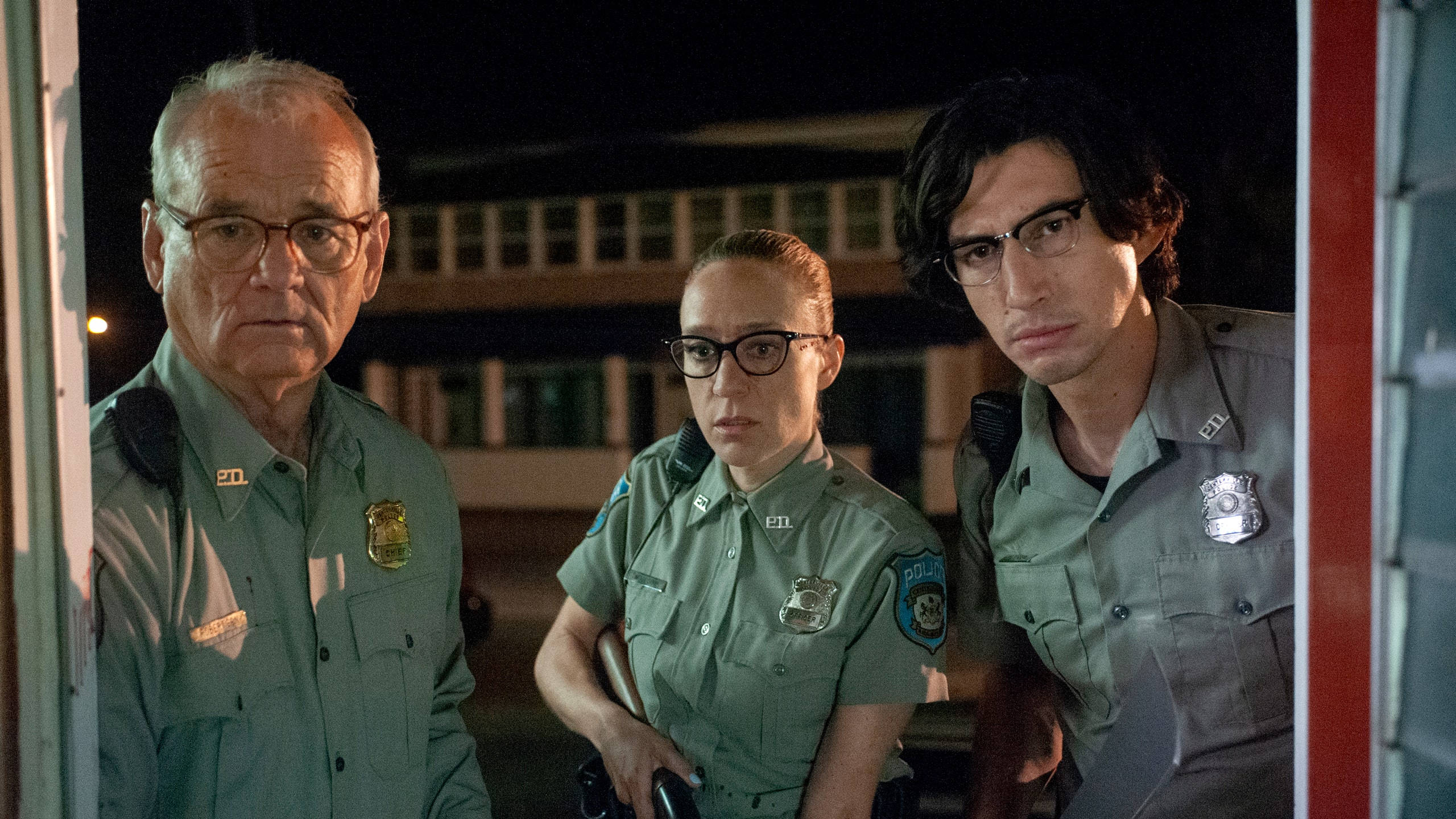 adam driver 桌布 壁紙 — 三名身穿綠色制服的急救人員在夜間警覺地站著，他們嚴肅的表情在黑暗建築背景下被溫暖的燈光照亮。