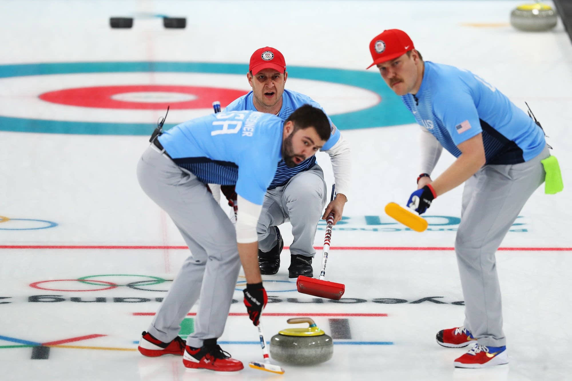 Olympics 桌布 壁紙 — 三名身穿藍色制服的男性冰壺運動員專注地，其中一人推著花崗岩石在冰上滑行，而隊友則在掃冰。
