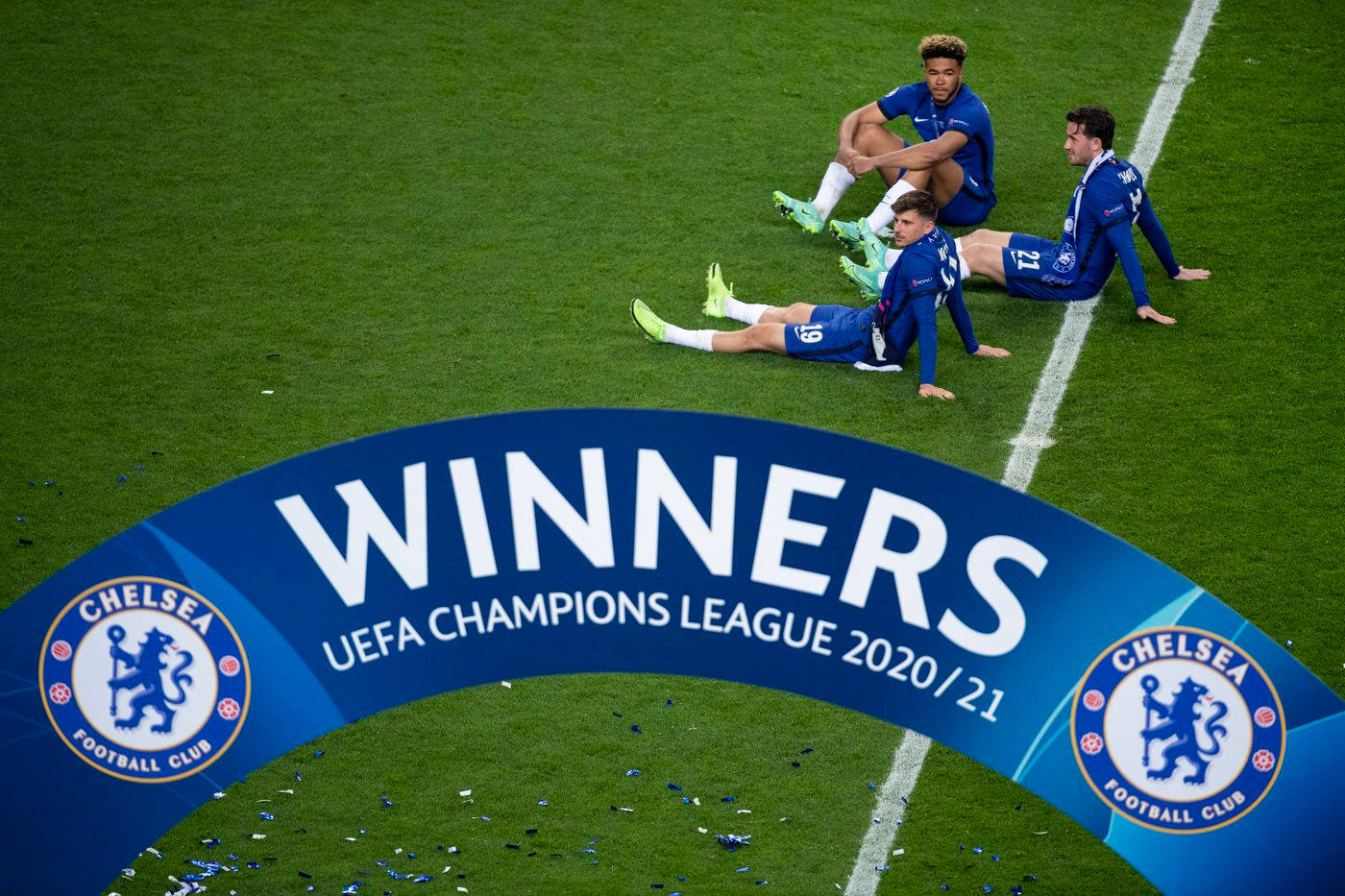 Champions League 桌布 壁紙 — 三名身穿藍色球衣的切爾西足球員疲憊地坐在綠色球場上，上方是'WINNERS UEFA CHAMPIONS LEAGUE 2020/21'拱門。