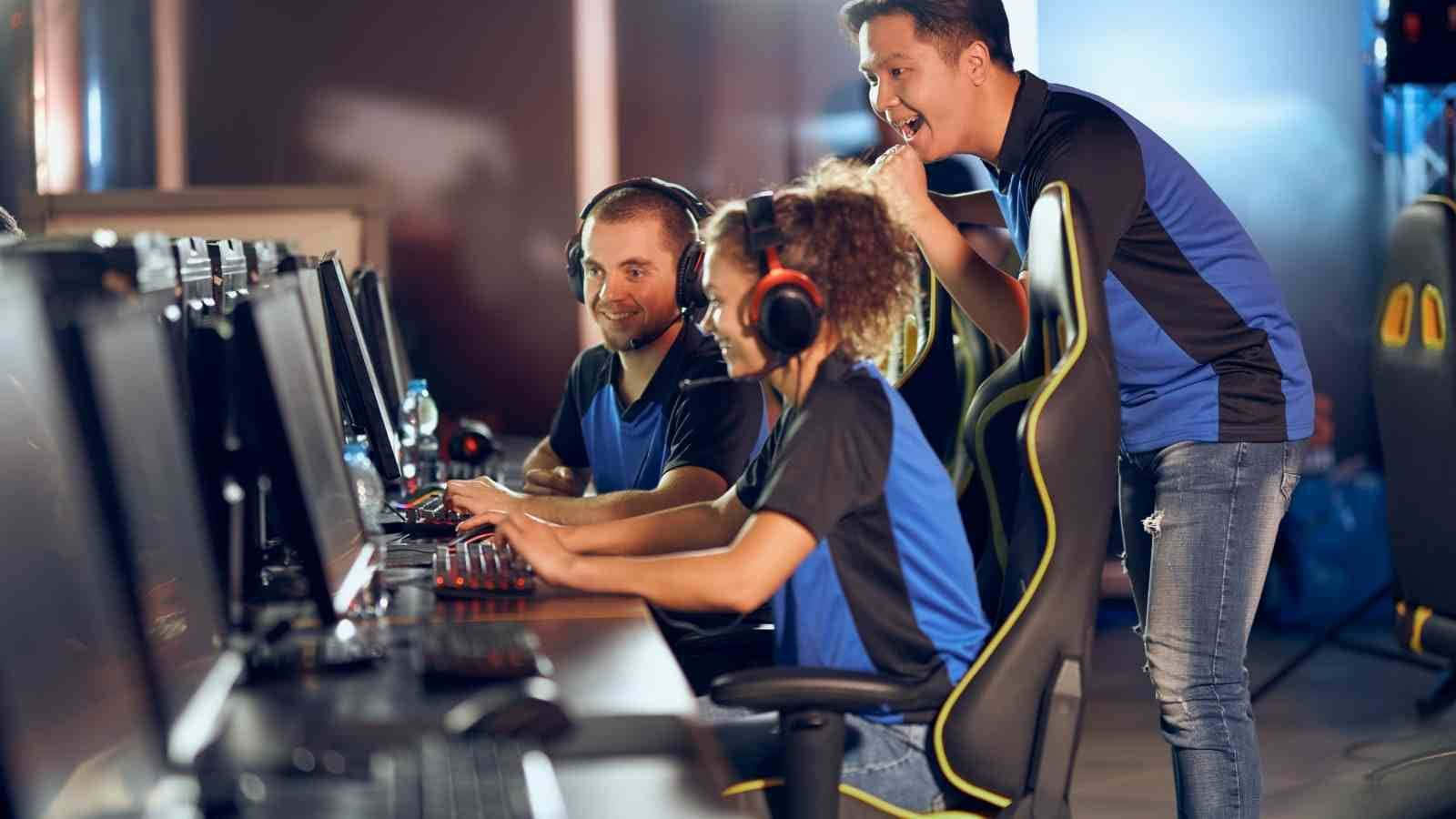 Esports 桌布 壁紙 — 三名身穿藍色球衣的玩家在燈光昏暗的電競場館中，伴隨著歡呼的興奮情緒，激烈地玩電腦。