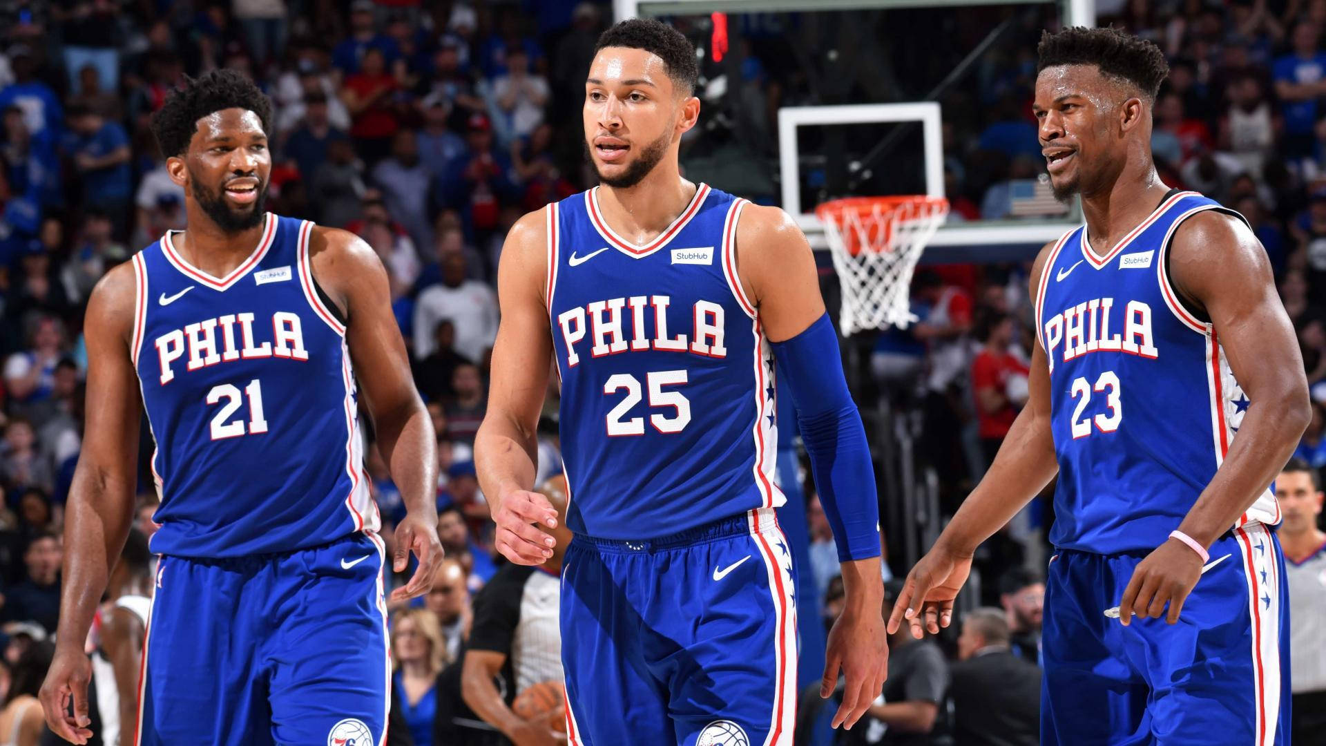 76ers 桌布 壁紙 — 三名身穿藍色球衣的費城76人籃球運動員在比賽期間一起走在燈光明亮的球場上。