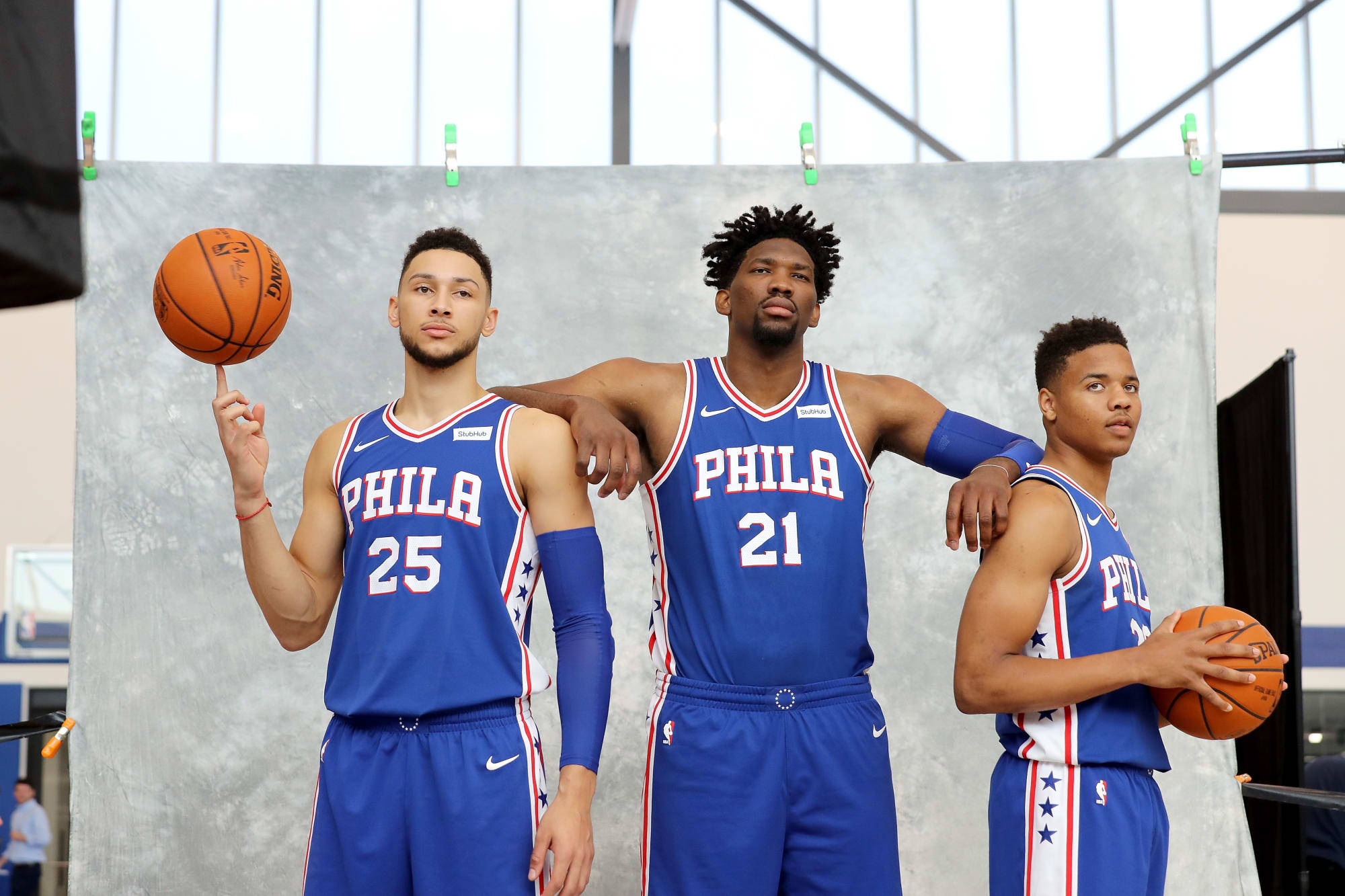 76ers 桌布 壁紙 — 三名身穿藍色球衣的費城76人籃球運動員在灰色背景下自信地拿著籃球擺姿勢。