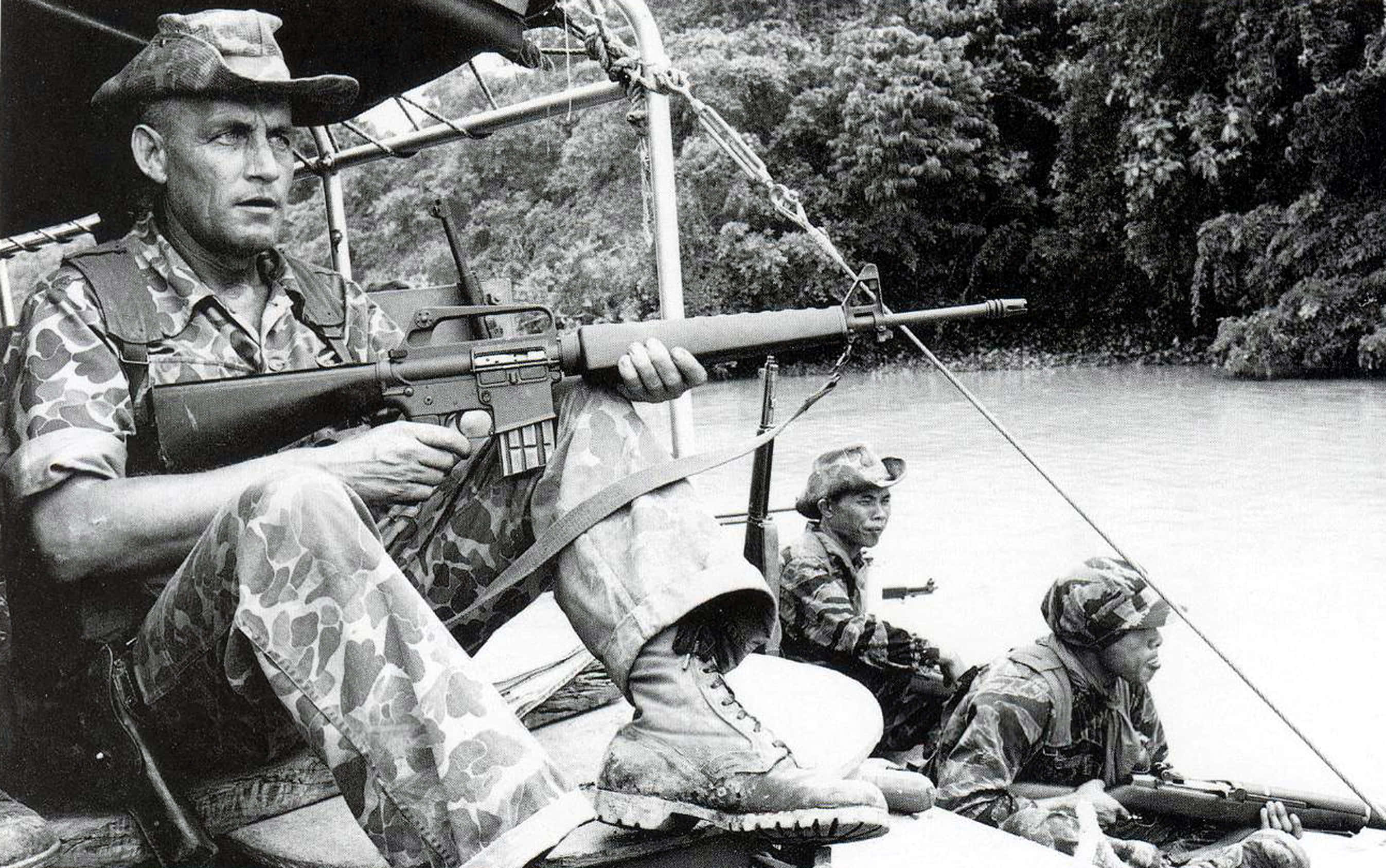 Vietnam 桌布 壁紙 — 三名身穿迷彩裝備的士兵警覺地坐在一艘船上，船在河流中，周圍是茂密的叢林樹葉。