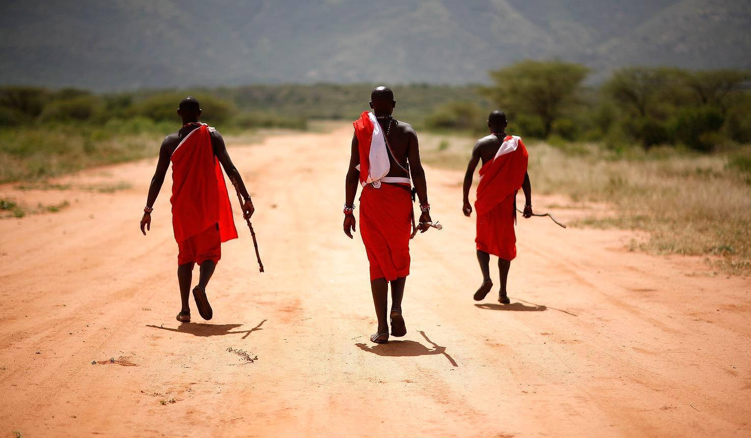 Kenya 桌布 壁紙 — 三名身穿鮮紅色Shukas的馬賽男子在烈日下沿著塵土飛揚的非洲道路走遠。