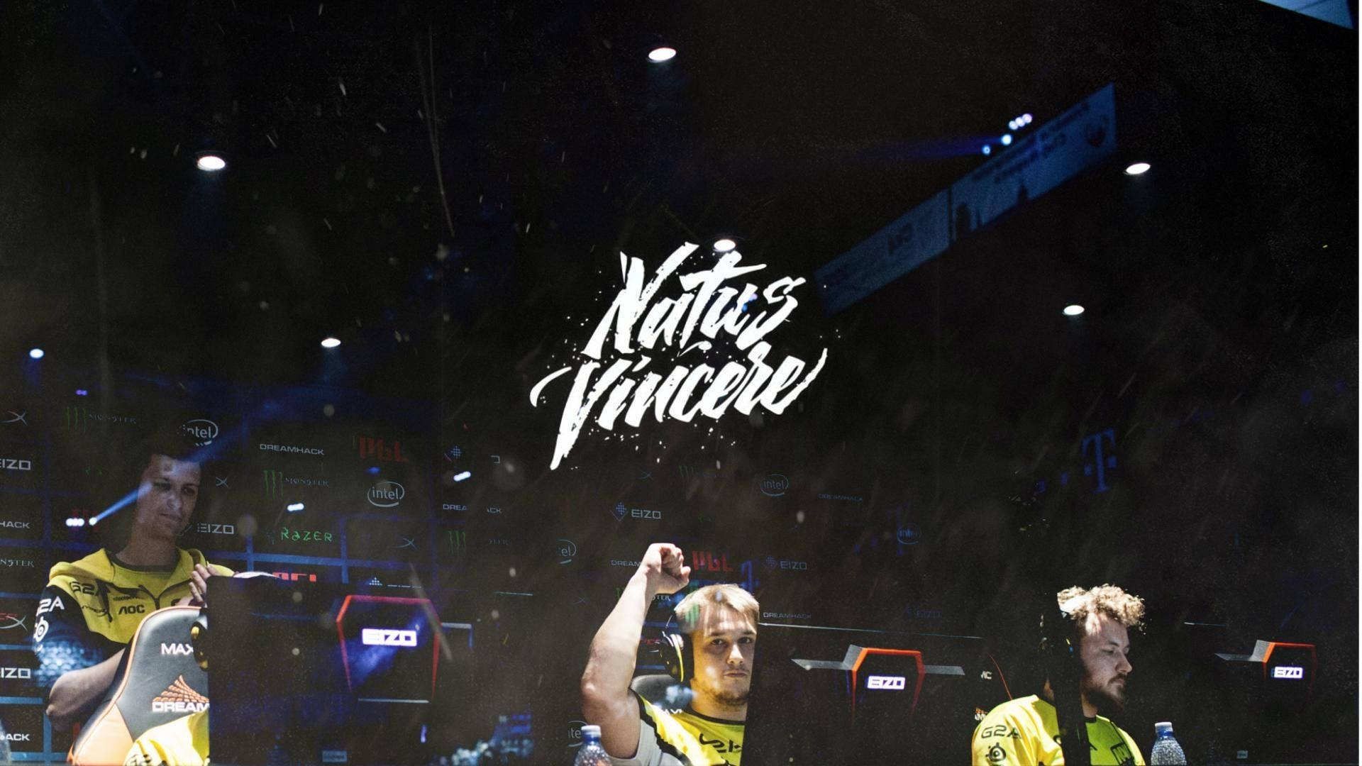 Natus Vincere 桌布 壁紙 — 三名身穿黃色球衣的電競選手在明亮的舞台燈光下慶祝勝利，『Natus Vincere』標誌在他們上方閃耀。