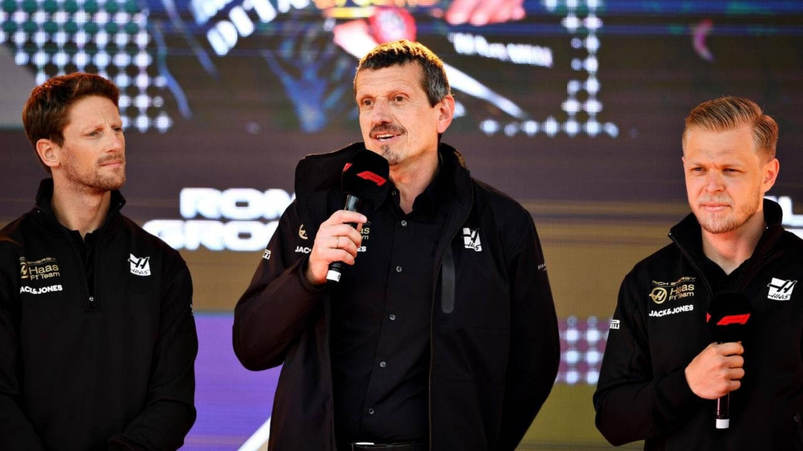 桌布 壁紙 — 三名身穿黑色Haas F1 Team外套的男子站在舞台上，其中一人對著麥克風說話，背景是發光的數位背景。