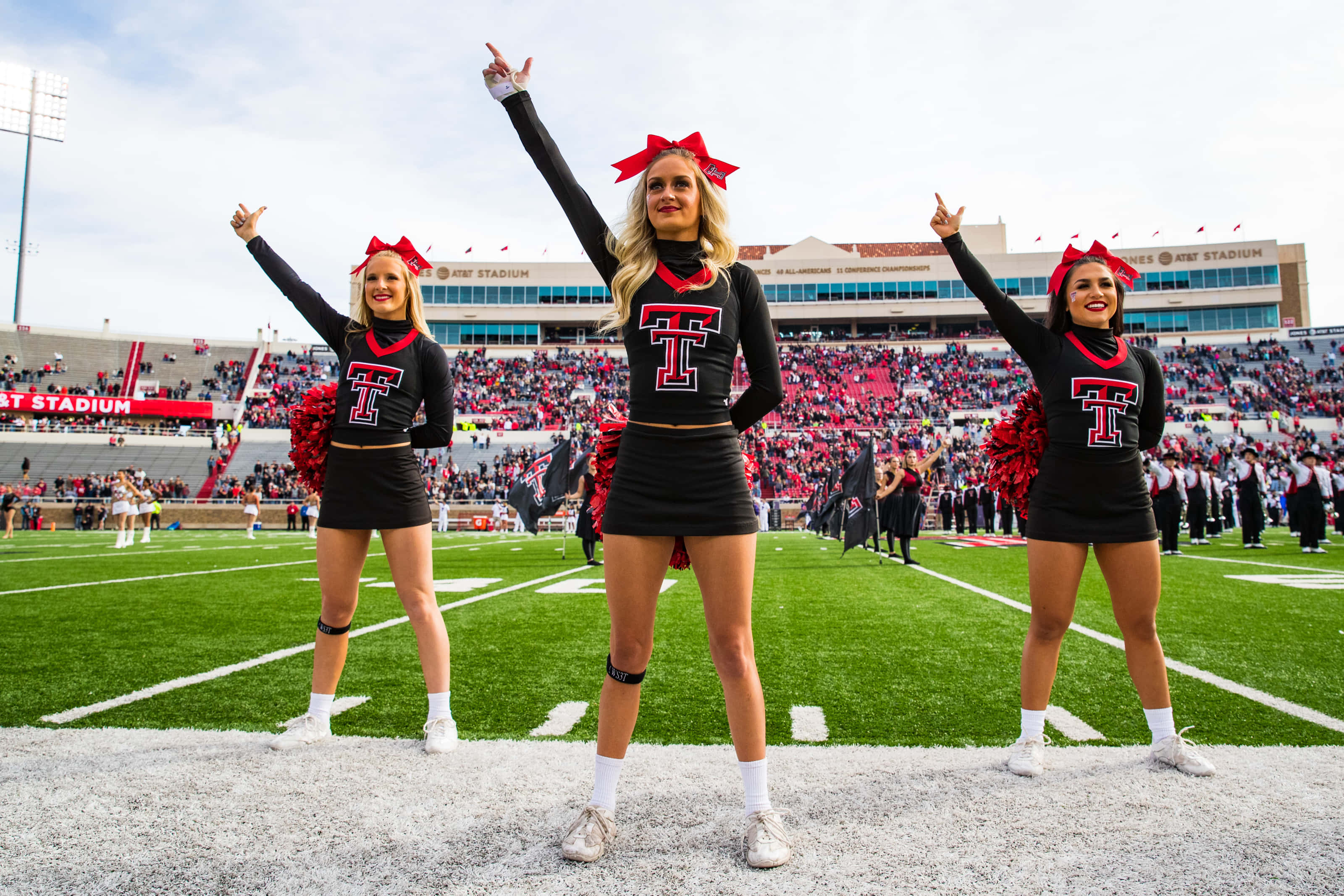 Texas Tech 桌布 壁紙 — 三名身穿黑色和紅色制服的啦啦隊員在一個擠滿觀眾的體育館後面的足球場上充滿活力地擺姿勢。