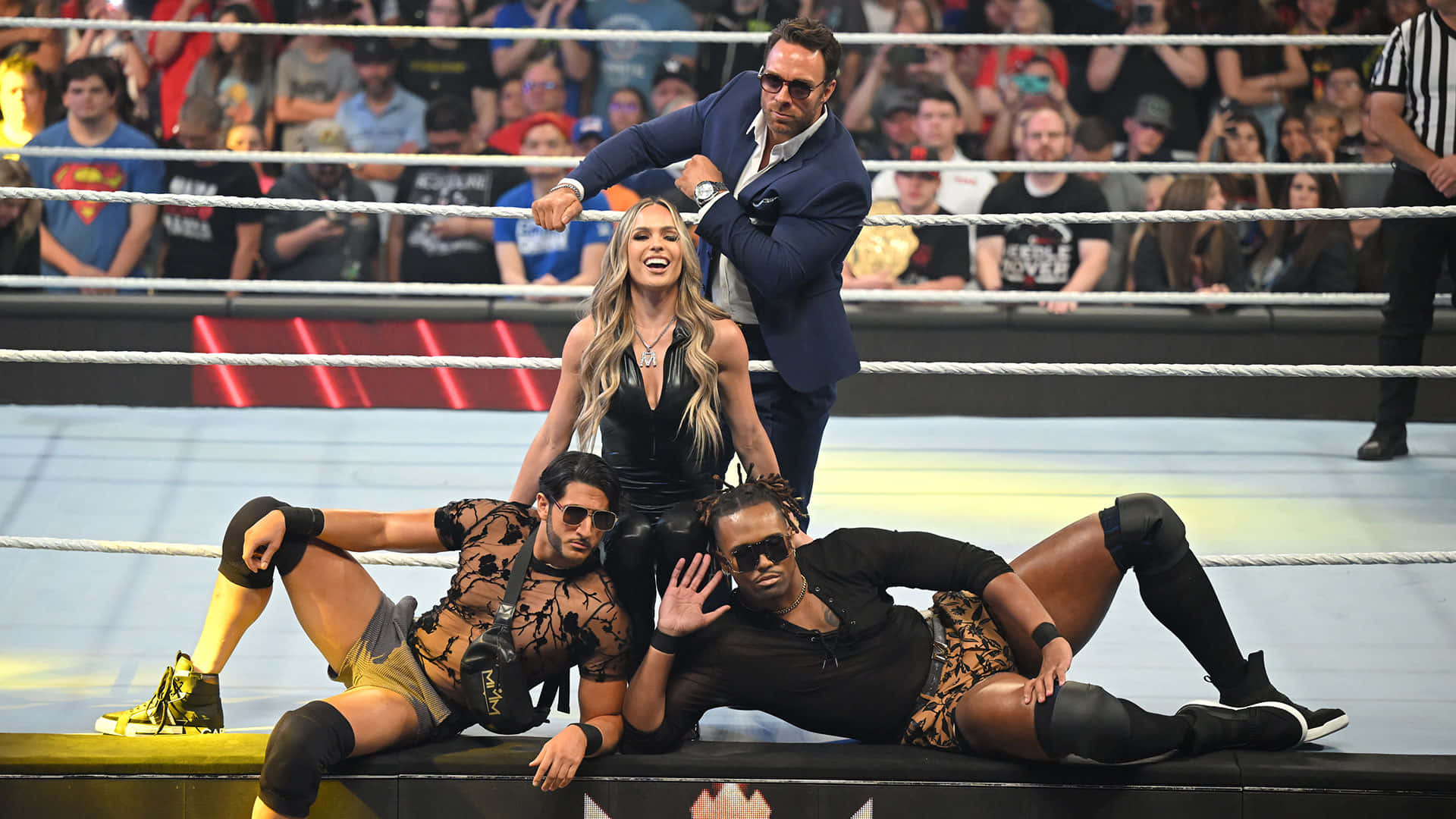 WWE 桌布 壁紙 — 三名身穿黑色服裝的摔角手在擂台邊休息，而一名穿西裝的男子在他們上方對著歡呼的人群做手勢。