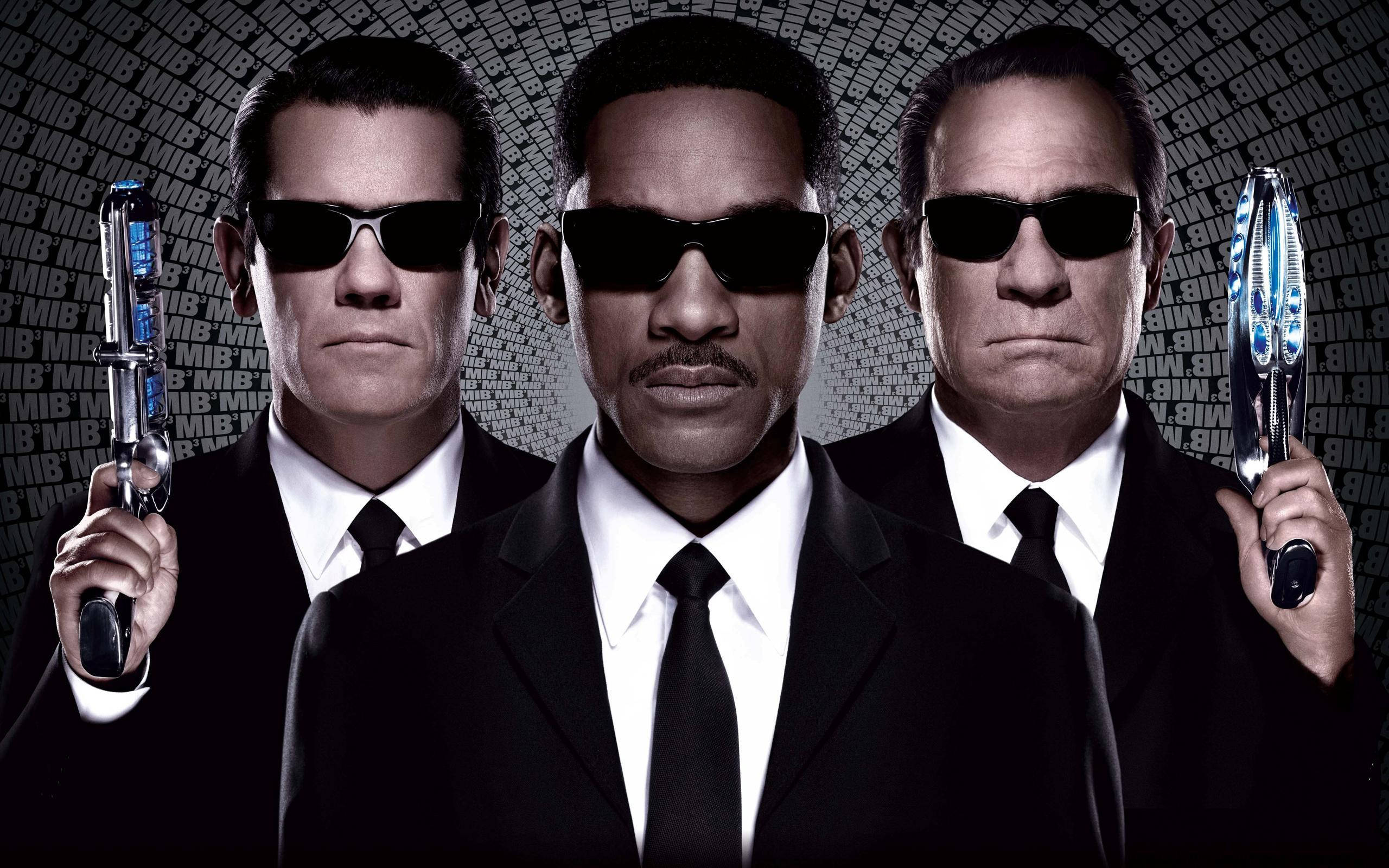 Men In Black 桌布 壁紙 — 三名身穿黑色西裝、戴著太陽眼鏡的男子，在重複的「MIB」圖案背景前手持未來武器。