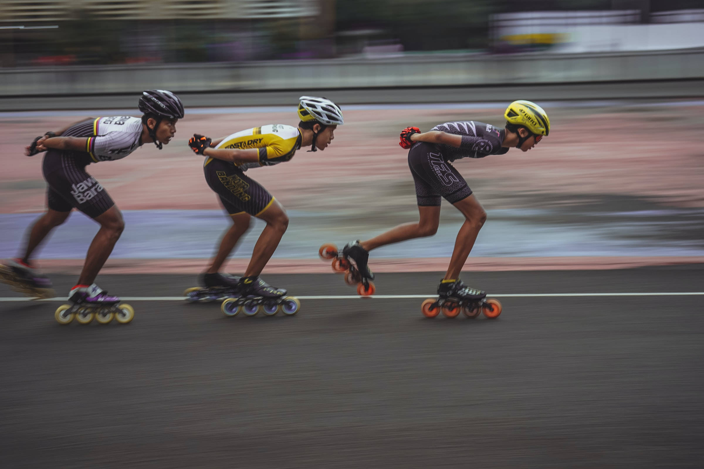 Athletic 桌布 壁紙 — 三名速度滑冰選手穿著賽車裝備，在室內賽道上激烈前傾比賽，背景呈現動態模糊效果。