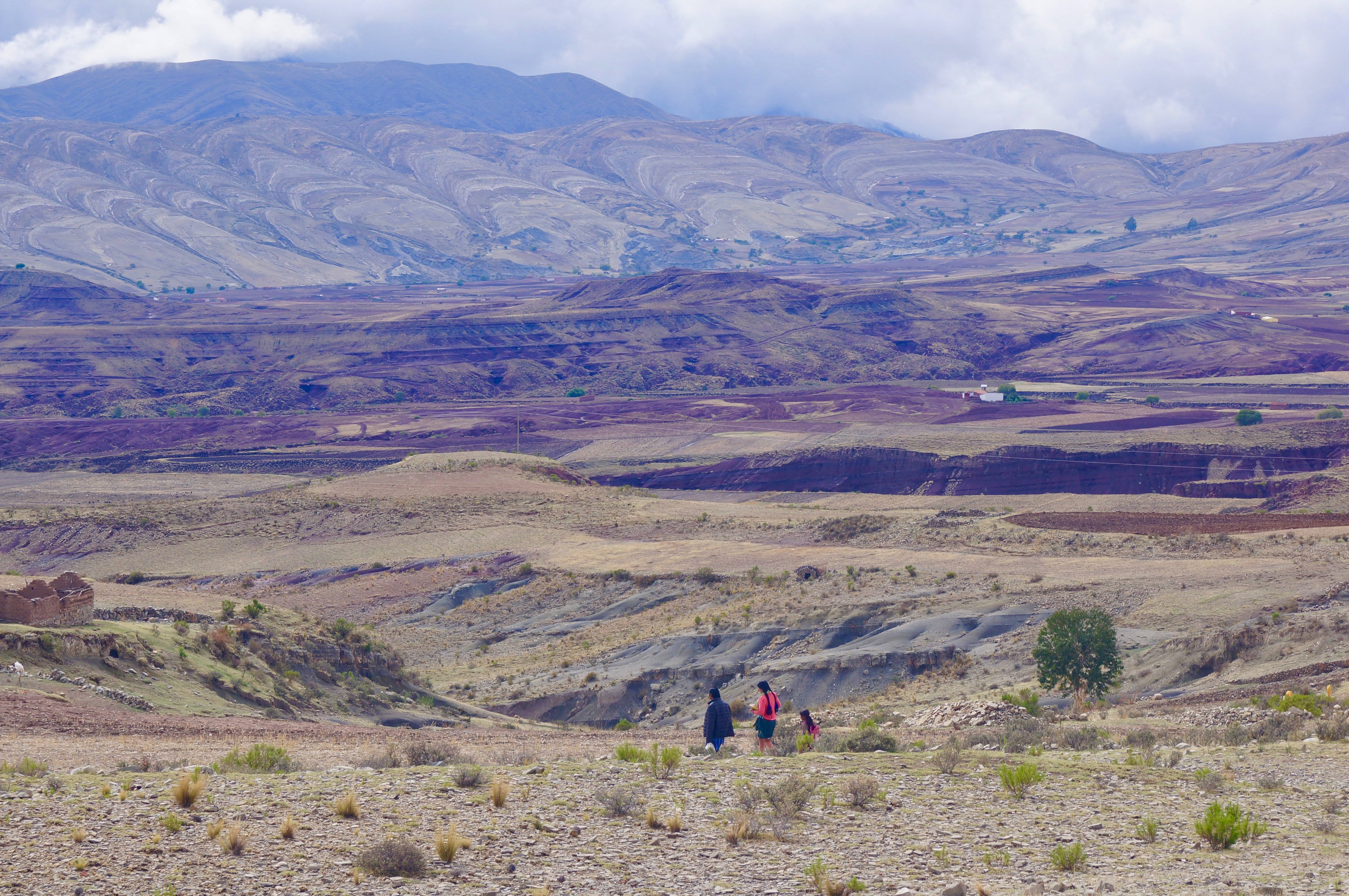 Bolivia 桌布 壁紙 — 三名遠足者穿越廣闊乾燥的山谷，山谷中有層層疊疊的紫色和赭色山丘，天空多雲。