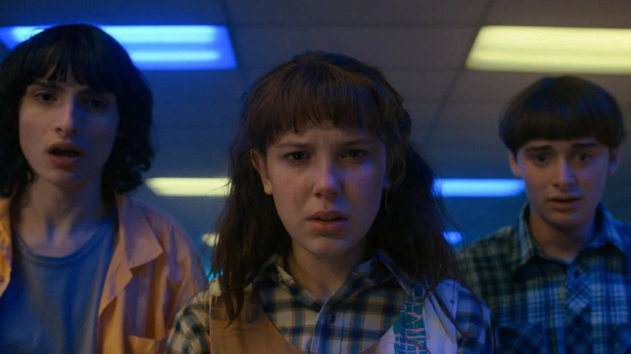 Stranger Things 4 桌布 壁紙 — 三名青少年在昏暗的房間裡，在藍色和黃色光芒的螢光燈下震驚地凝視。