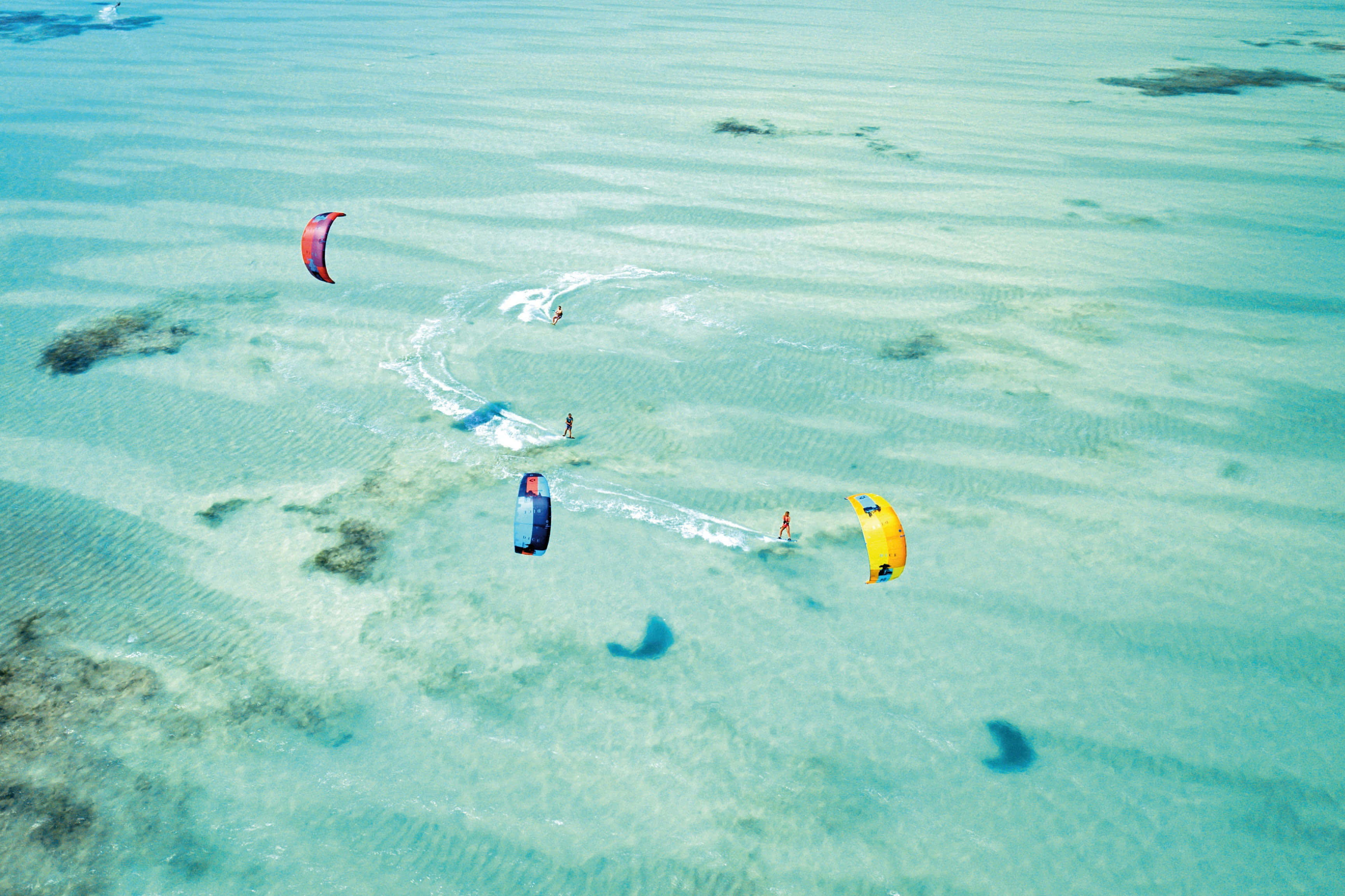 Beach Water Sports 桌布 壁紙 — 三名風箏衝浪者在碧綠淺灘上滑行，身後留下白色痕跡的鳥瞰圖。