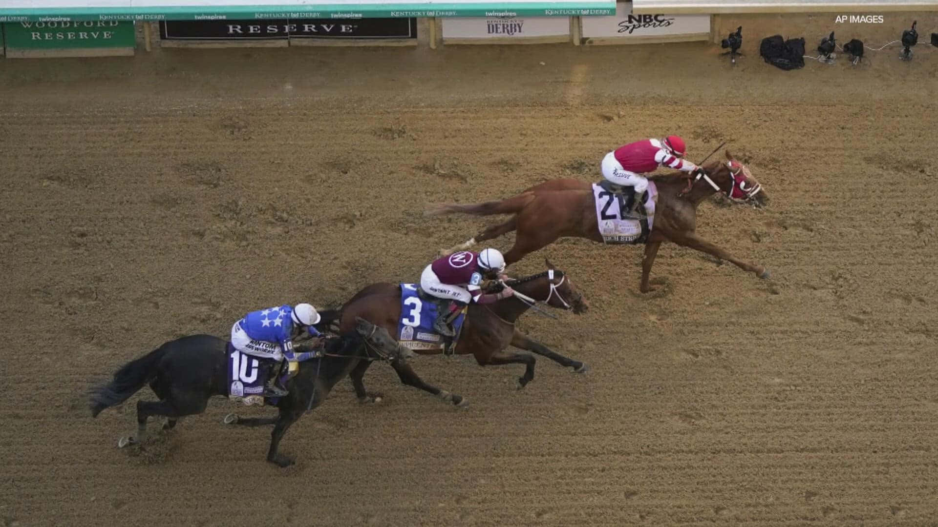 Kentucky Derby 桌布 壁紙 — 三名騎師身穿彩色絲綢，在泥地賽道上策馬奔馳，背景有體育館橫幅。