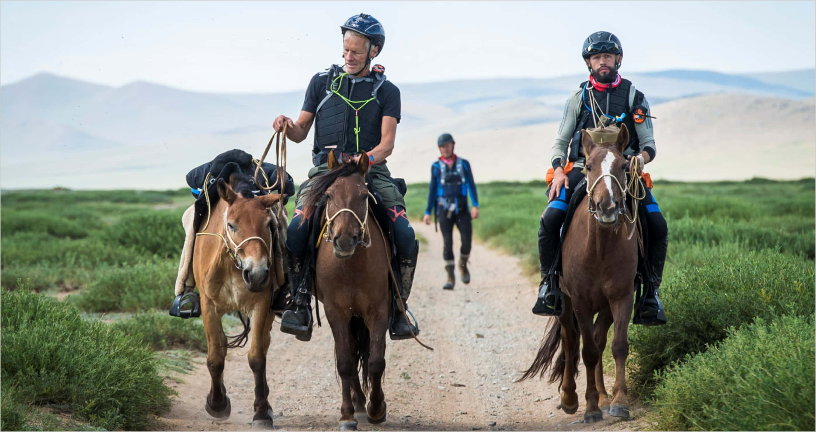 Mongolia 桌布 壁紙 — 三名騎手騎著馬，沿著一條塵土飛揚的小徑，穿過廣闊的綠色草原，遠處有山脈，天空明亮。