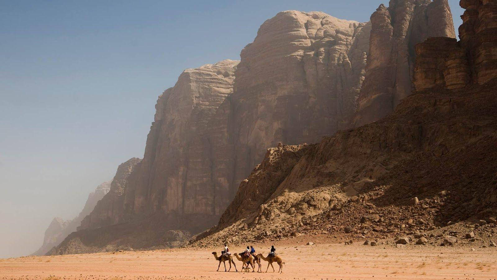 Jordan 桌布 壁紙 — 三名騎手騎著駱駝穿越廣闊的沙漠山谷，下方是高聳的砂岩懸崖，上方是清澈的藍天。