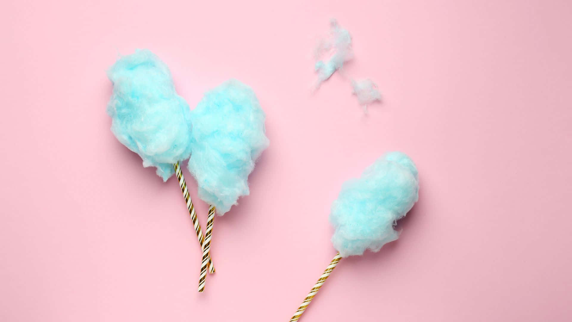 Cotton Candy 桌布 壁紙 — 三團蓬鬆藍色棉花糖插在條紋棒上，置於柔和的粉紅色背景上，周圍散落著糖絲。