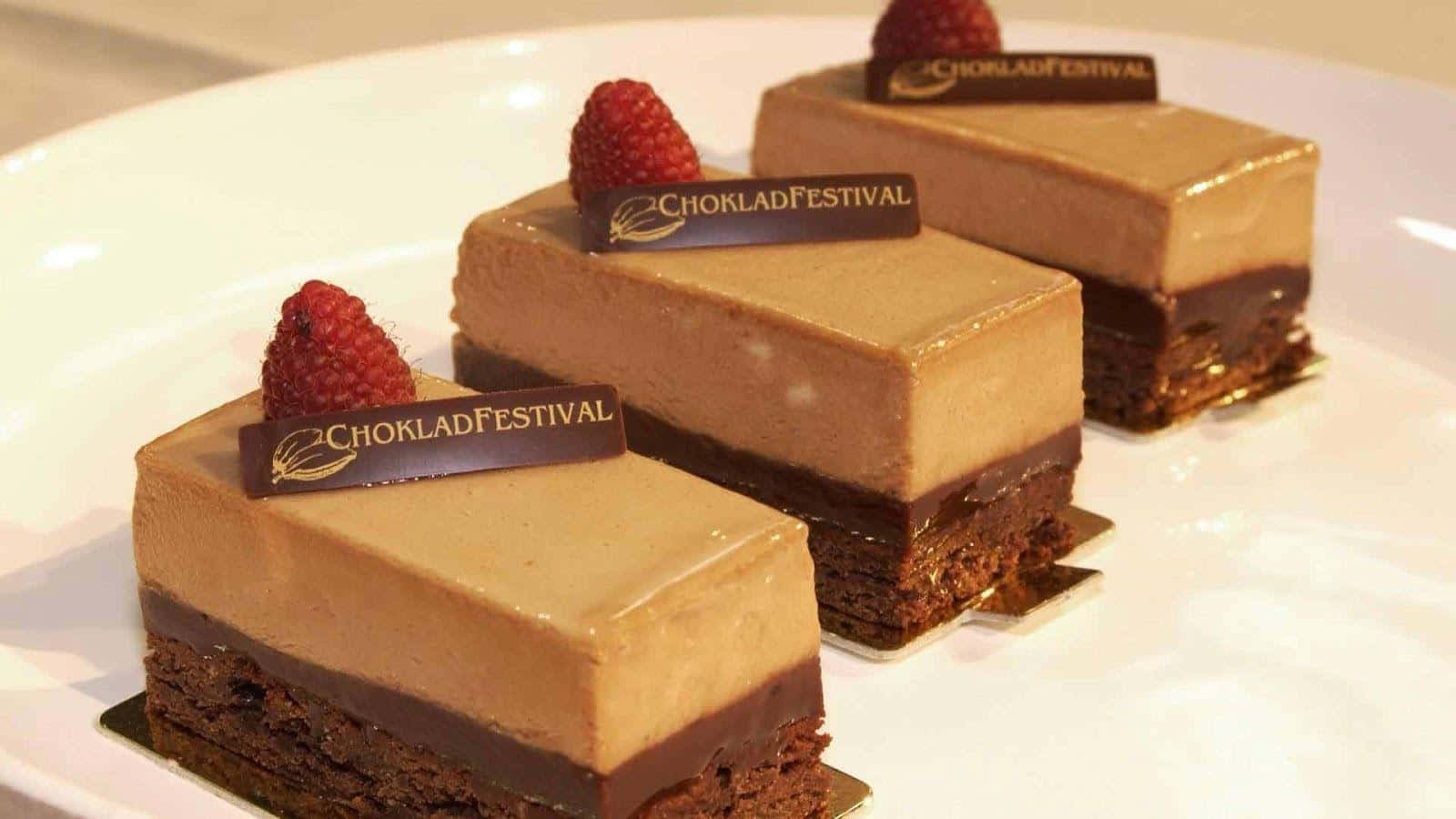 Chocolate Cake 桌布 壁紙 — 三塊亮面巧克力慕斯蛋糕，上面放有覆盆子和品牌標籤，優雅地擺放在白色盤子上。