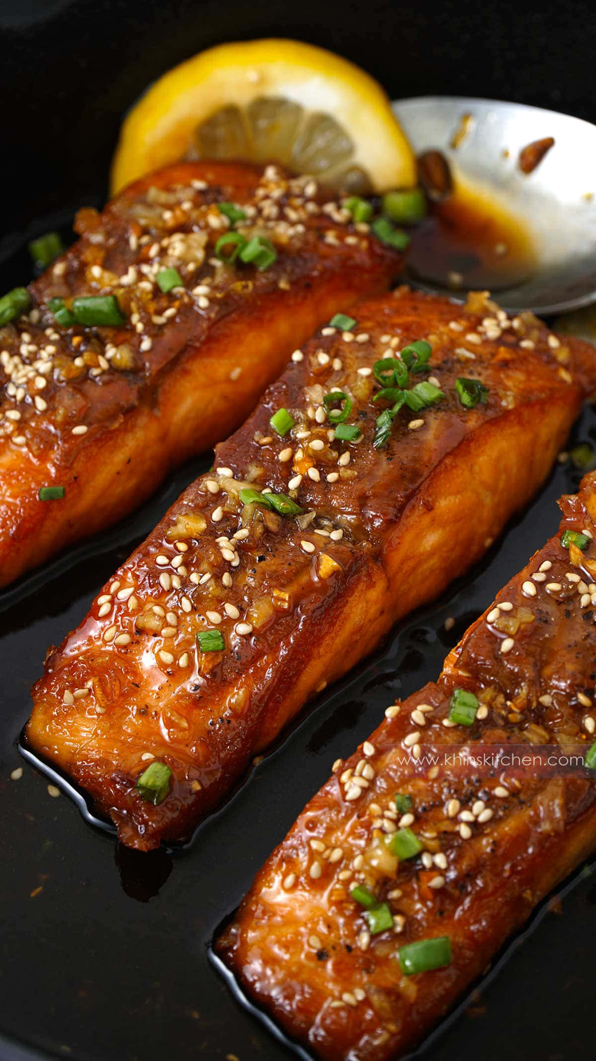 Salmon 桌布 壁紙 — 三塊塗上醬汁的鮭魚排，上面撒上芝麻和青蔥，放在一個深色平底鍋裡，旁邊放著一塊檸檬角。