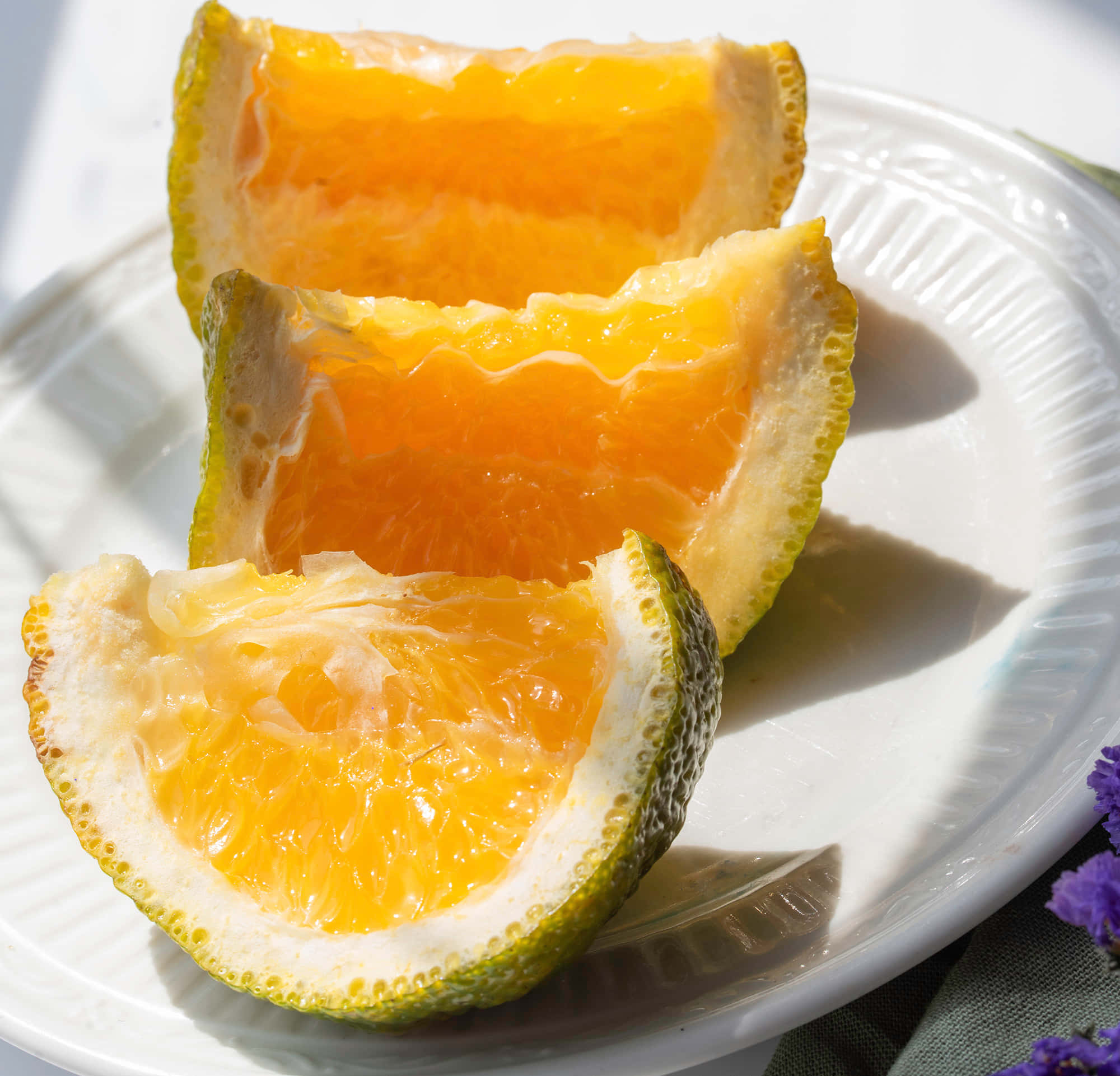 Fruits 桌布 壁紙 — 三塊多汁的橙瓣在白色盤子上，旁邊有紫色花朵，在明亮陽光下閃閃發光。