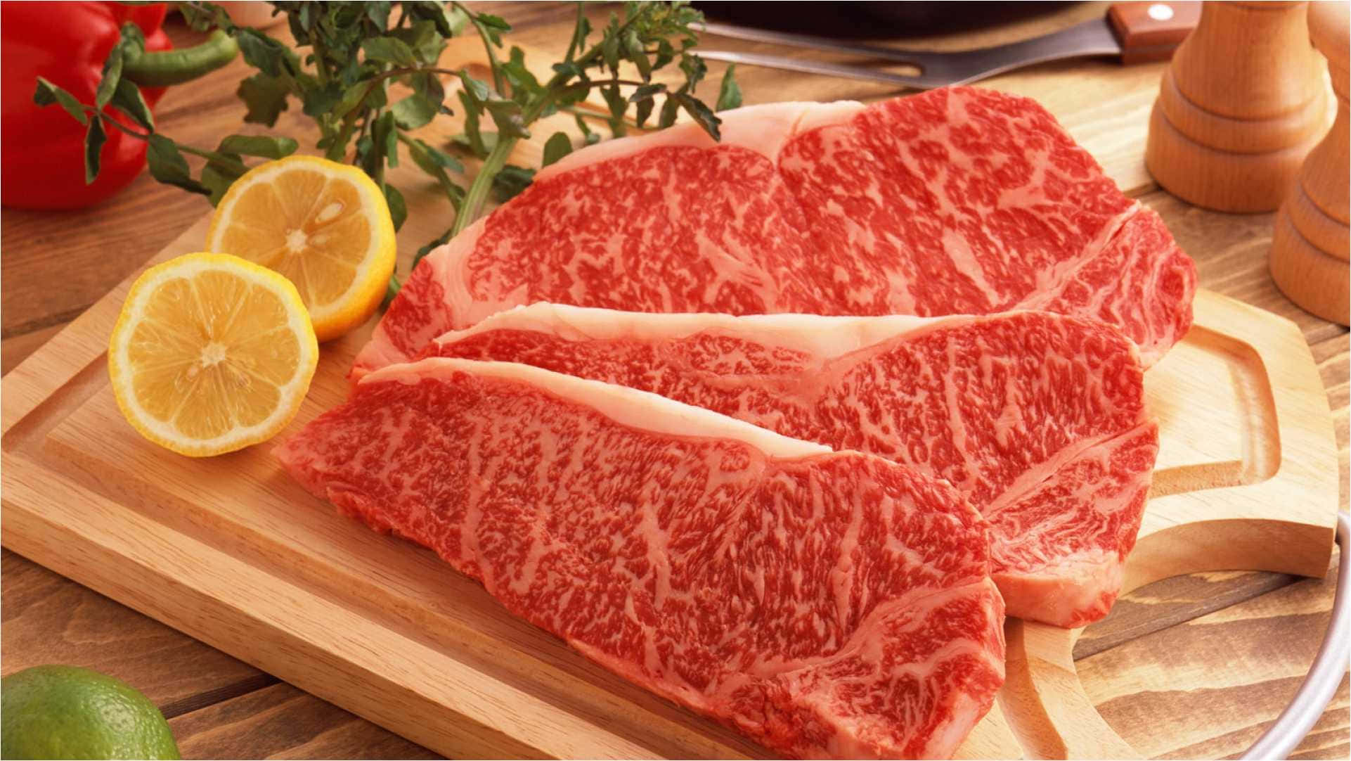 meat 桌布 壁紙 — 三塊大理石紋牛排放在木砧板上，旁邊有檸檬片和新鮮香草。