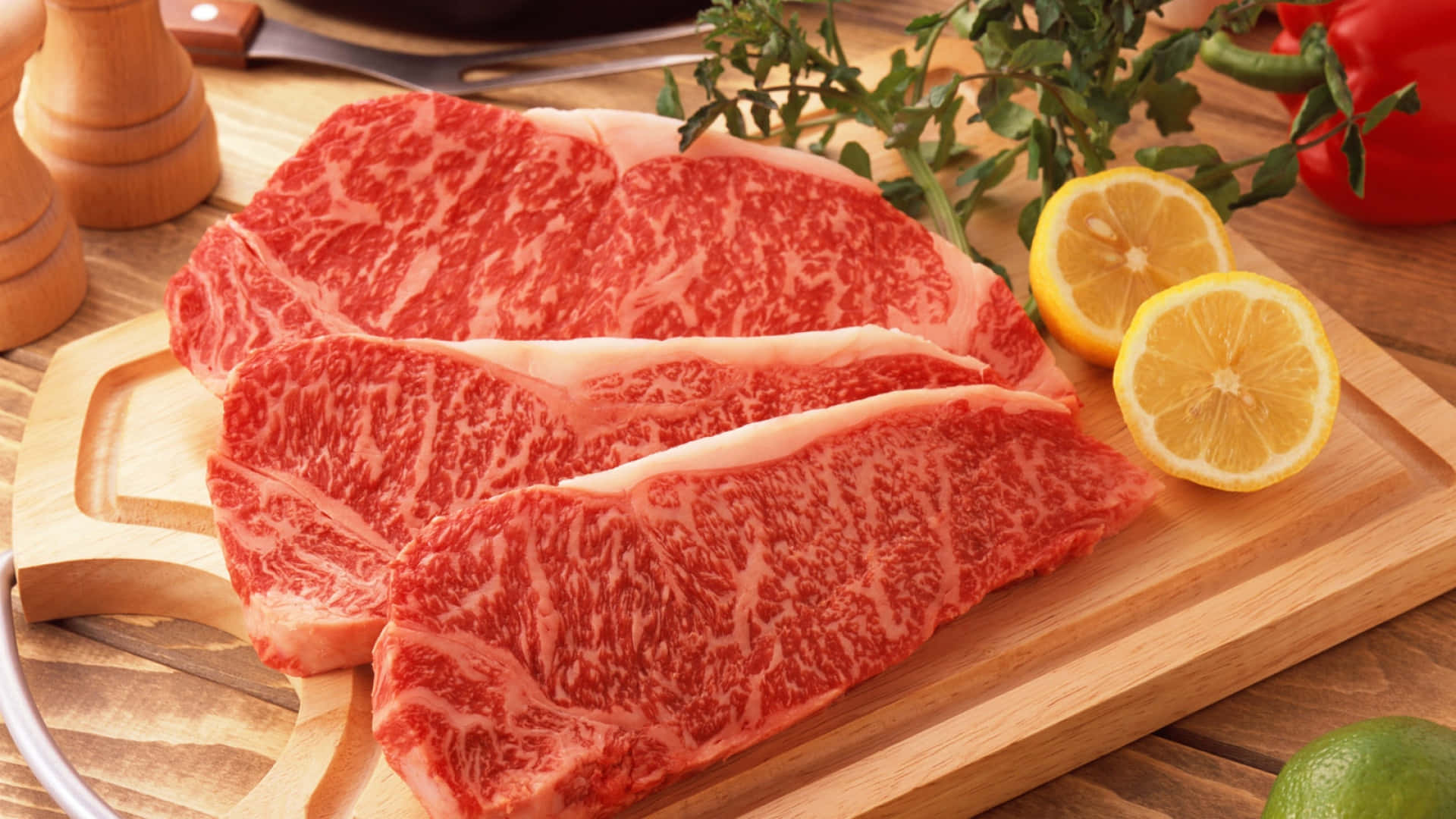 Red Meat 桌布 壁紙 — 三塊大理石紋牛排放在木製砧板上，旁邊有檸檬片和新鮮香草。