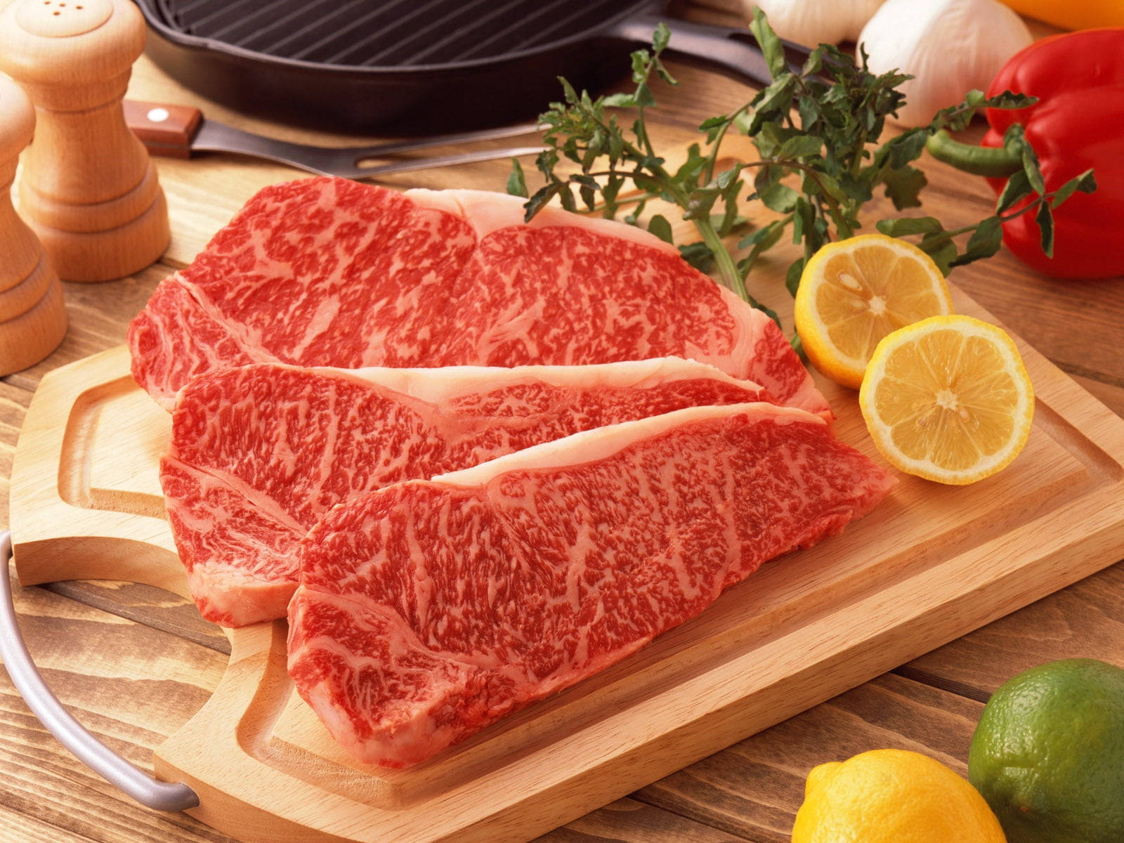meat 桌布 壁紙 — 三塊大理石紋牛排放置在木製砧板上，周圍有柑橘類香草和烹飪工具。