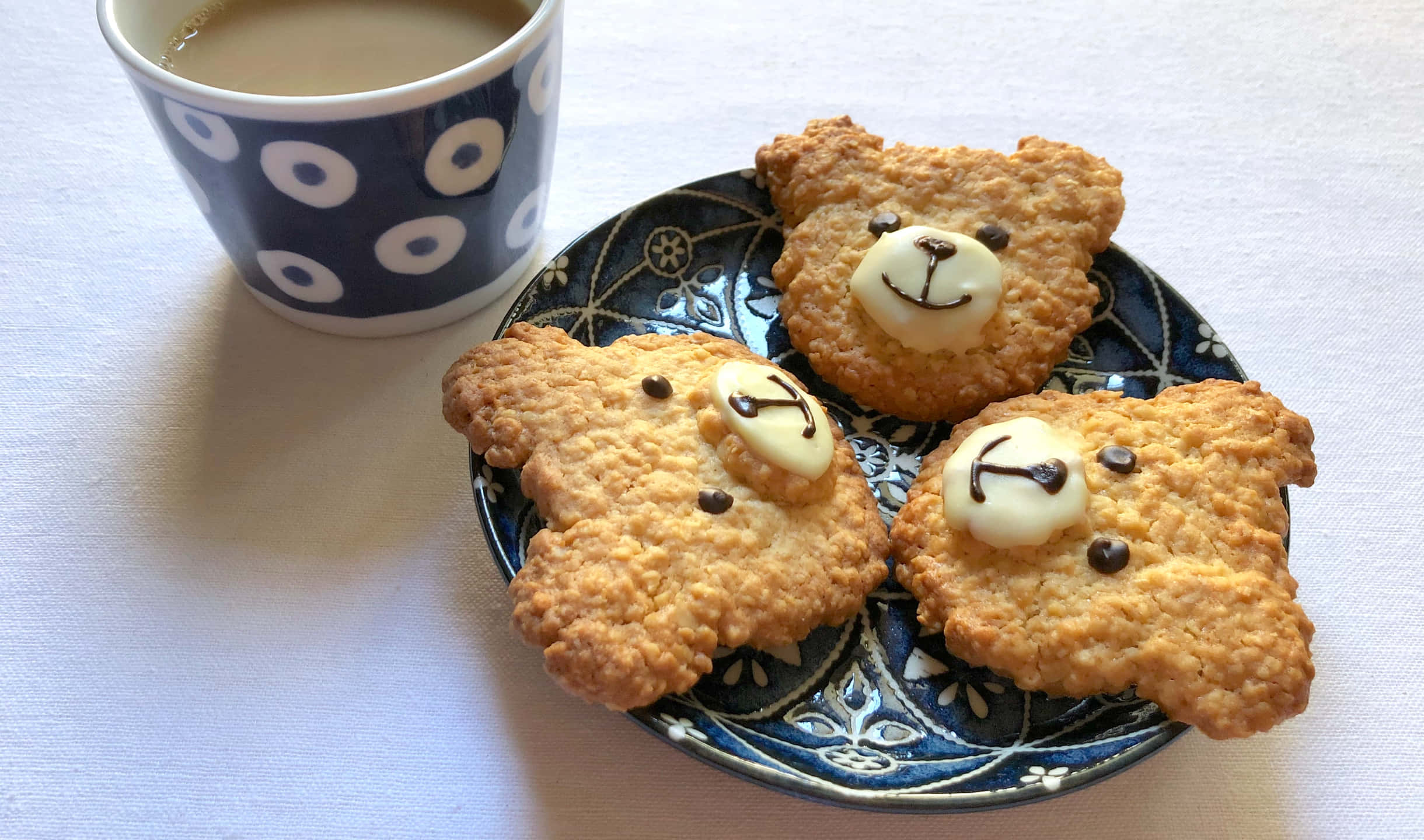 Teddy Bear 桌布 壁紙 — 三塊小熊形狀的燕麥餅乾，有巧克力眼睛和白色糖霜微笑，放在一個藍色圖案盤子上，旁邊有一杯圓點咖啡。