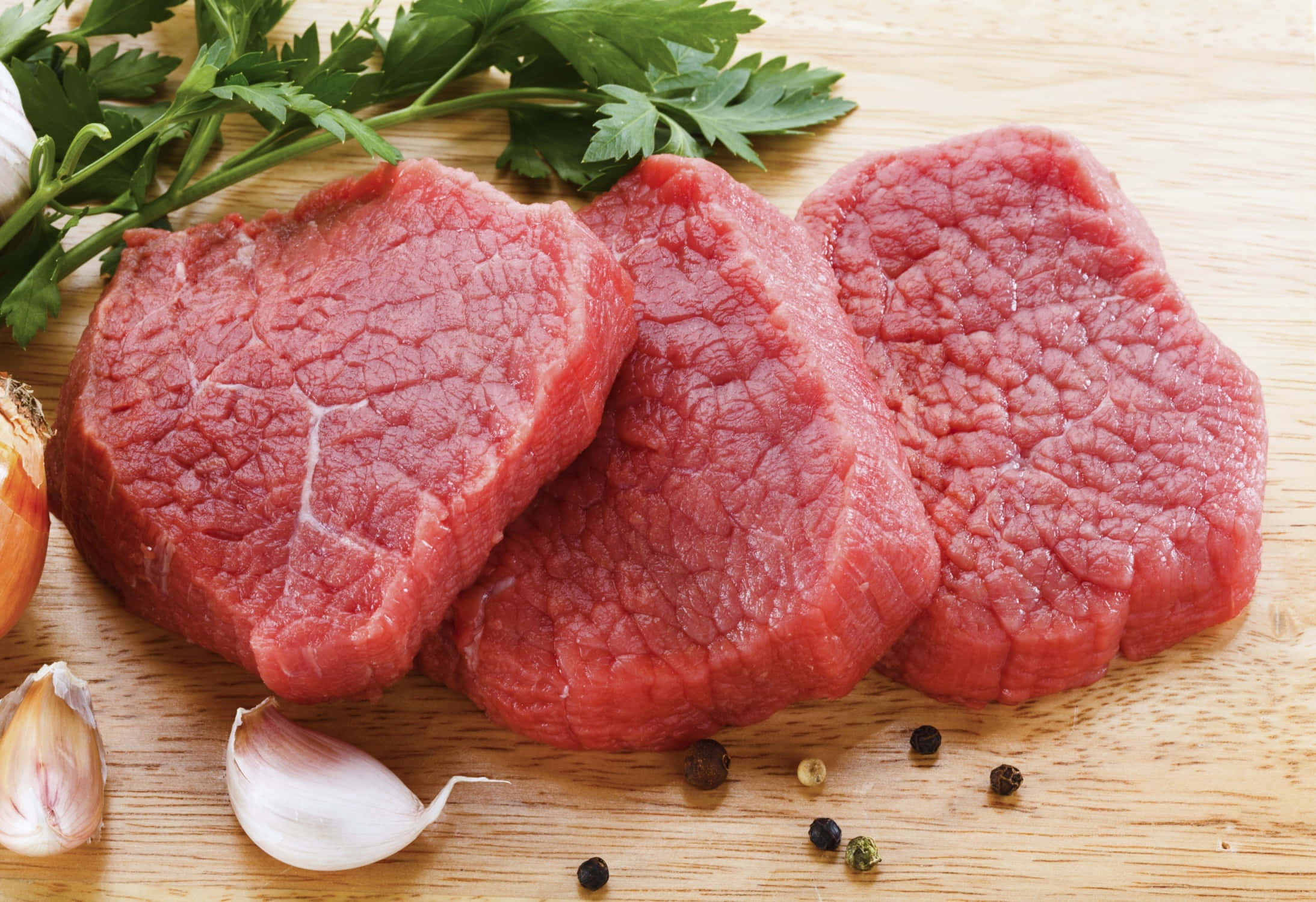 meat 桌布 壁紙 — 三塊帶有明顯大理石花紋的生牛肉排放在木板上，旁邊是新鮮的歐芹、大蒜和胡椒粒。
