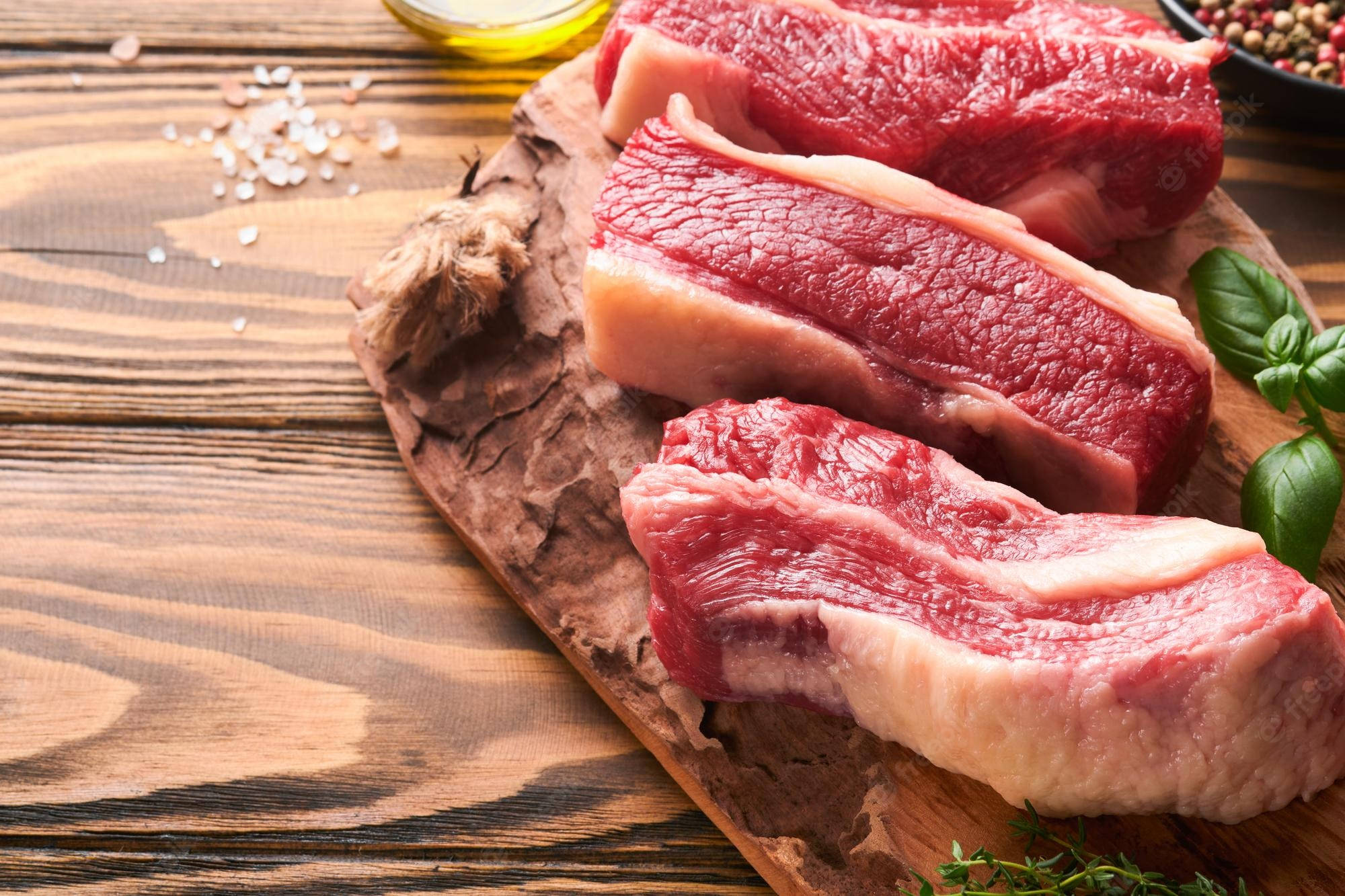 meat 桌布 壁紙 — 三塊帶有油花的生牛肉排放在一個鄉村風格的木砧板上，旁邊有鹽、油和新鮮香草。