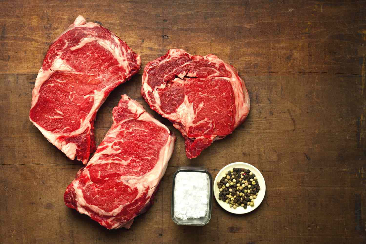 Ribeye Steak 桌布 壁紙 — 三塊帶有油花紋理的生牛肉排放在一塊鄉村風格的木板上，旁邊有鹽和胡椒粒。