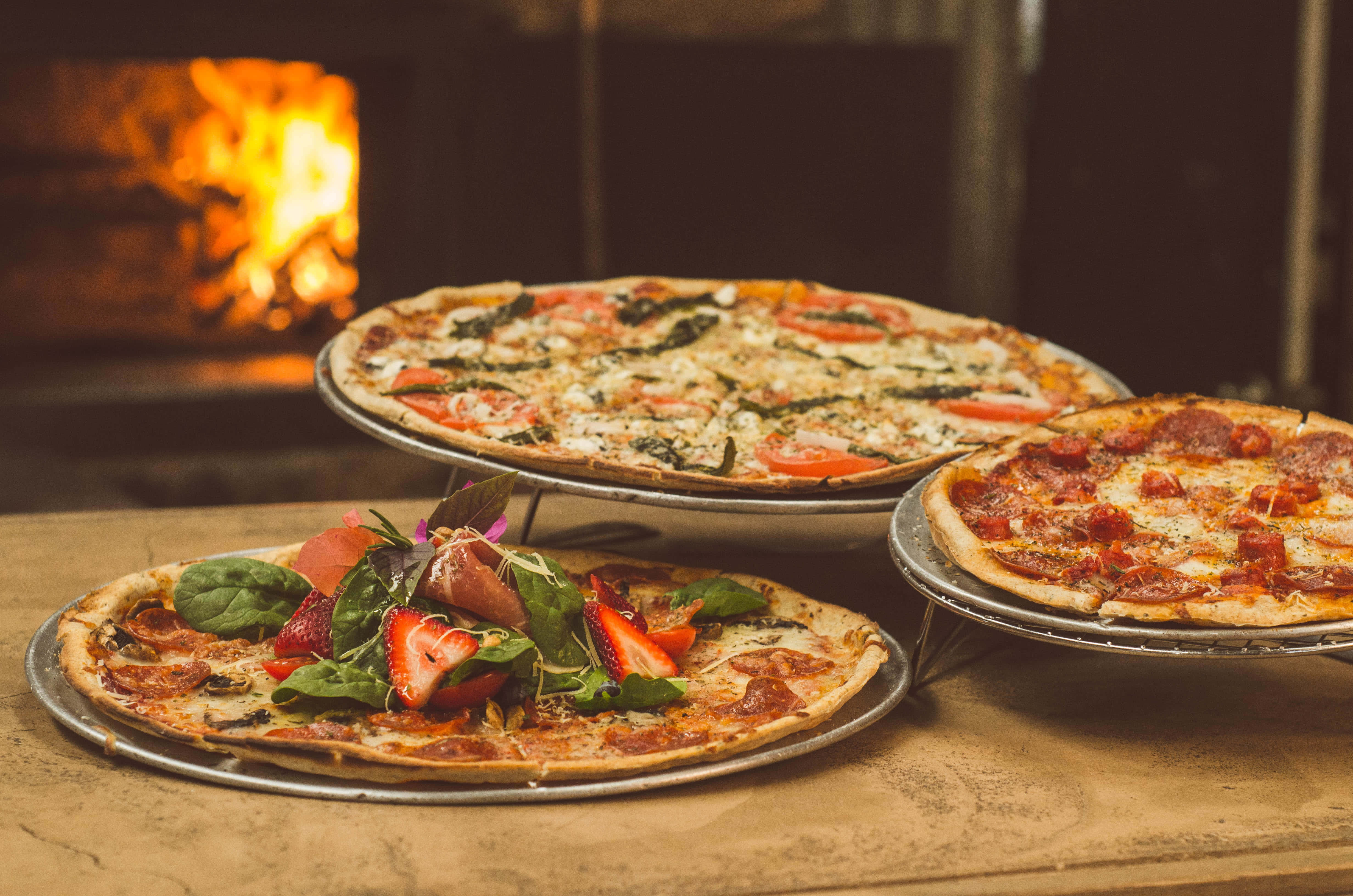 Pizza 桌布 壁紙 — 三塊手工披薩配上新鮮配料，放在金屬托盤上，置於鄉村風披薩店中發光的柴火烤爐前。