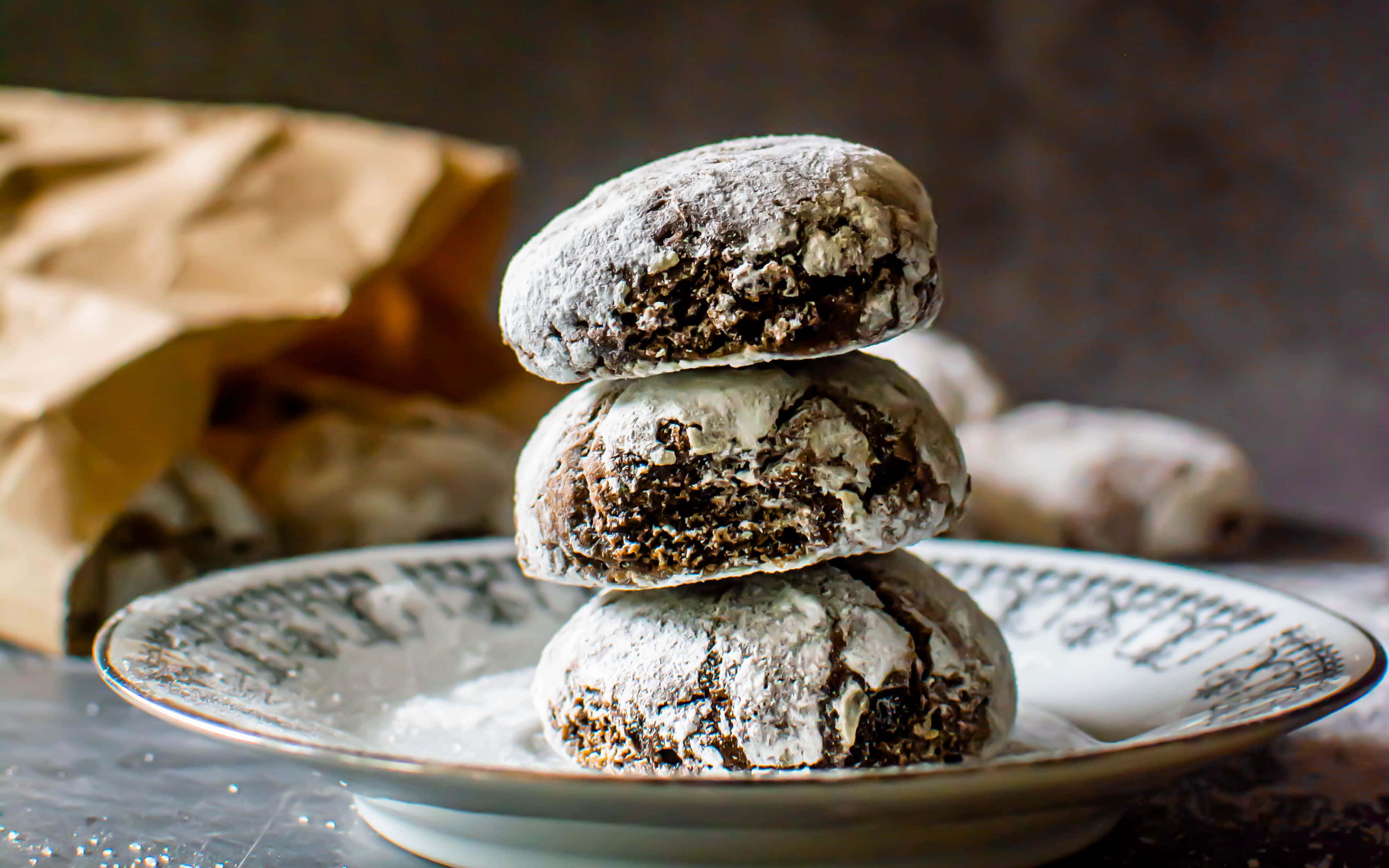 Cookies 桌布 壁紙 — 三塊撒上糖粉的巧克力餅乾疊放在有圖案的盤子上，背景有一個棕色的紙袋，呈現柔和焦點。