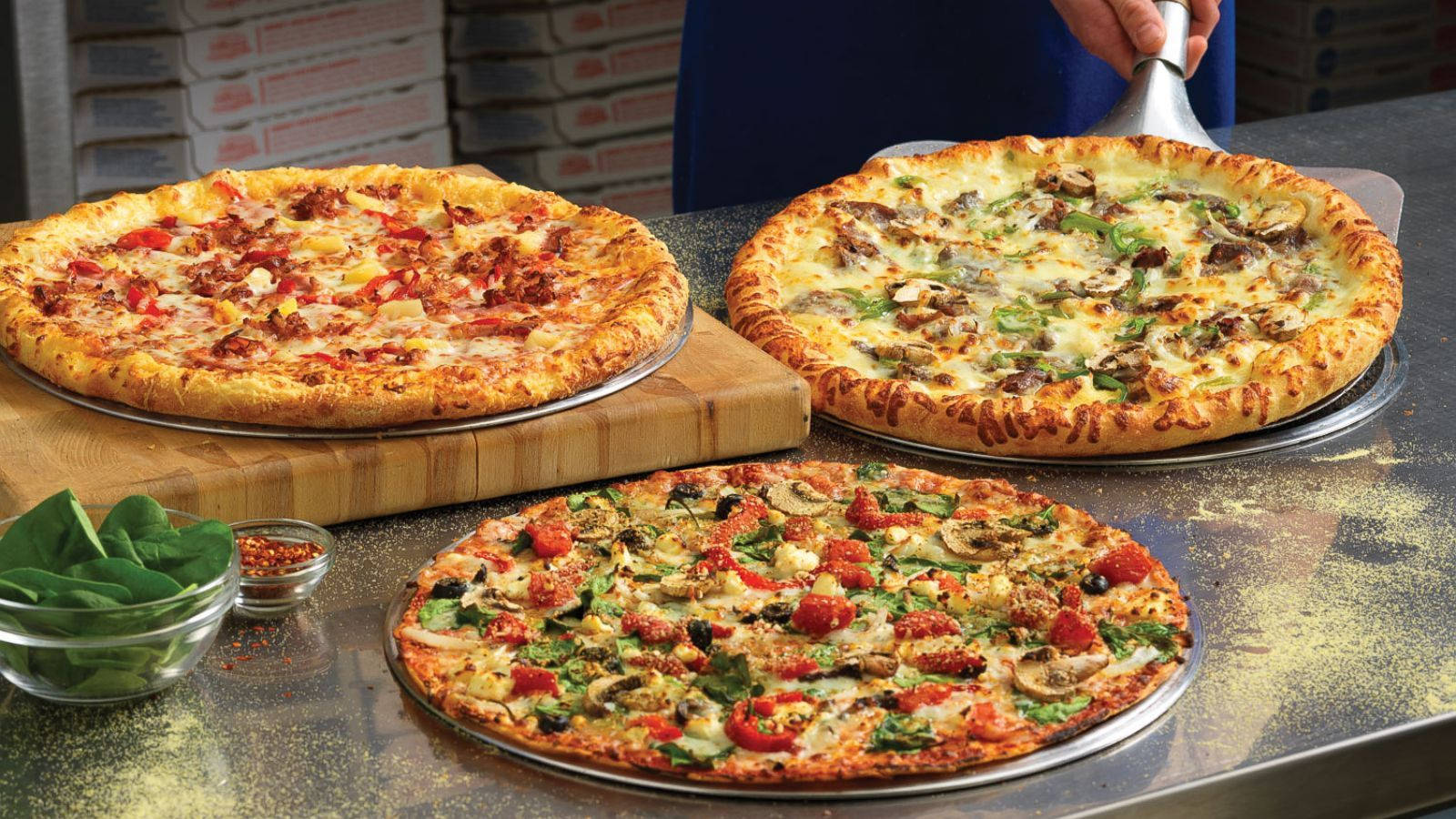 Dominos Pizza 桌布 壁紙 — 三塊新鮮出爐的披薩，上面有鮮豔的配料，放在繁忙披薩店廚房的金屬托盤上。