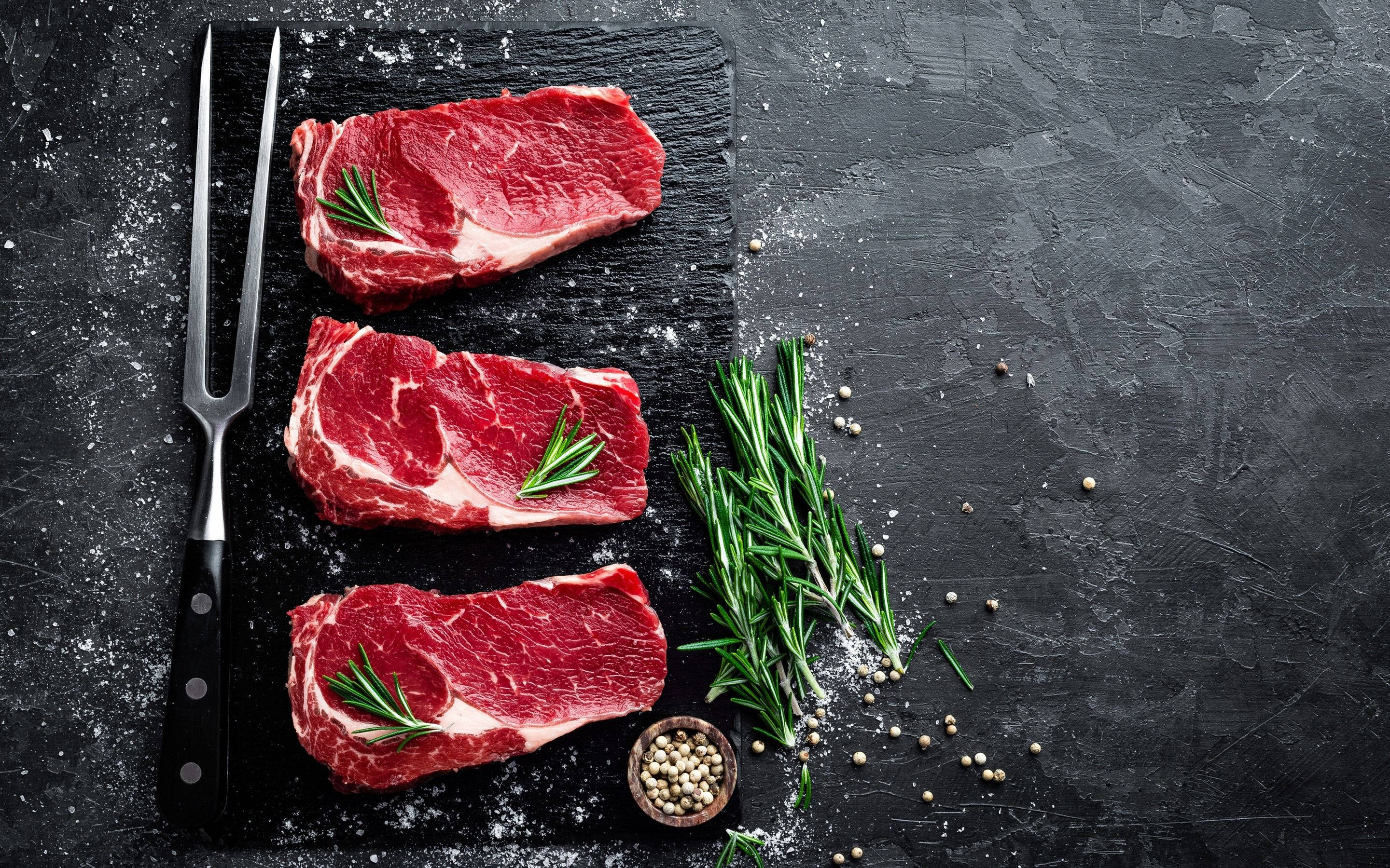 meat 桌布 壁紙 — 三塊生牛肉排以迷迭香點綴，放在深色石板板上，配有鹽、胡椒和一把切肉叉。