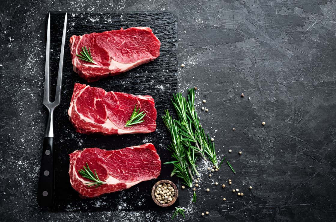 Red Meat 桌布 壁紙 — 三塊生牛肉排以迷迭香點綴，放在深色石板板上，旁邊有叉子和胡椒粒。