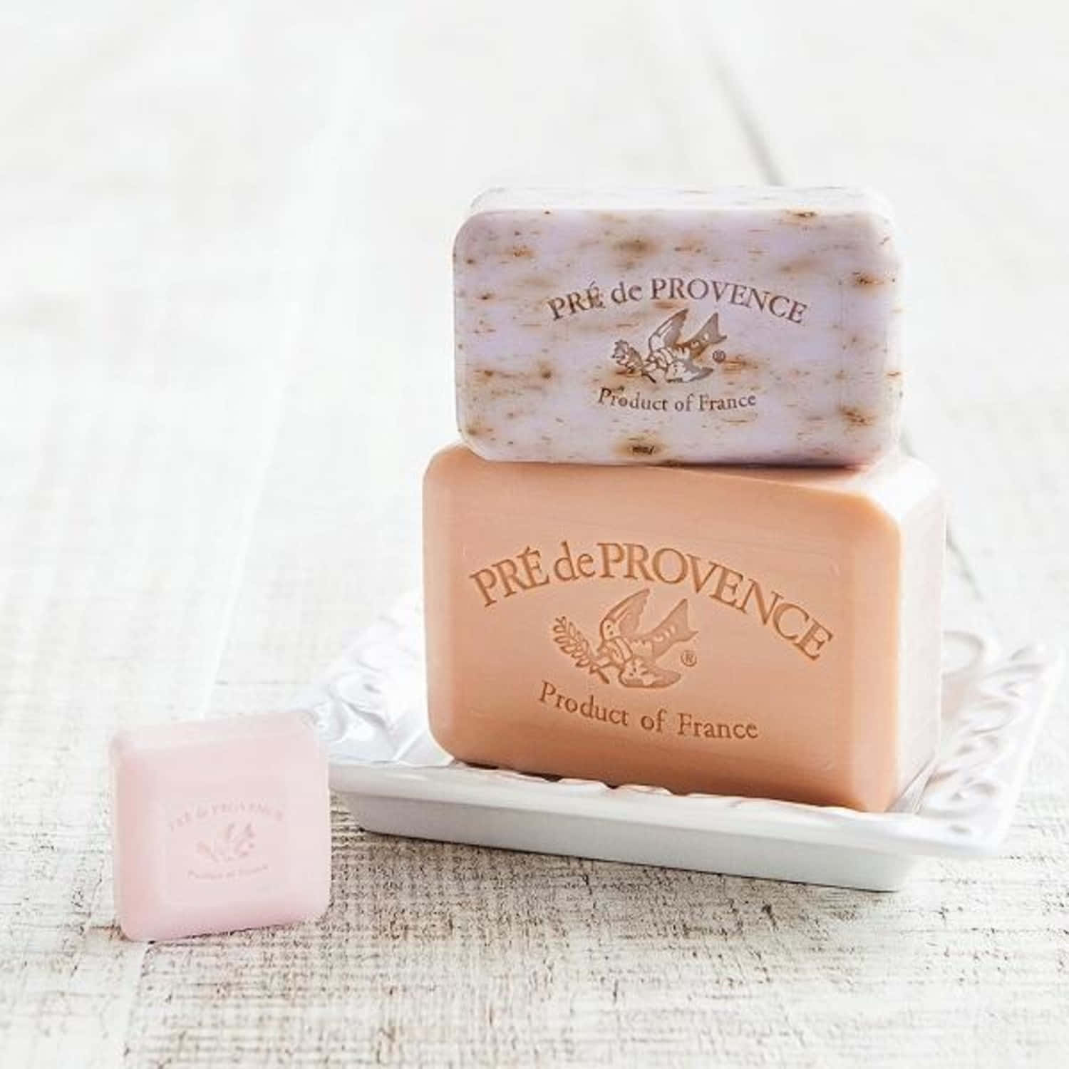 Square 桌布 壁紙 — 三塊疊放的PRE de PROVENCE肥皂，有蜜桃、薰衣草和粉紅色，放在一個白色陶瓷碟上，置於經風化的木質表面。
