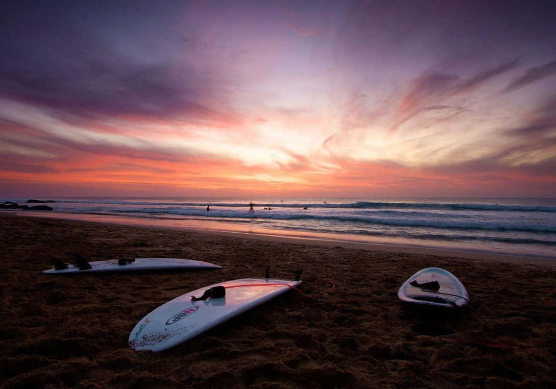 surfing 桌布 壁紙 — 三塊衝浪板擱在沙灘上，紫色、橙色和粉紅色的鮮豔雲彩在海洋上空的日落天空中蔓延。