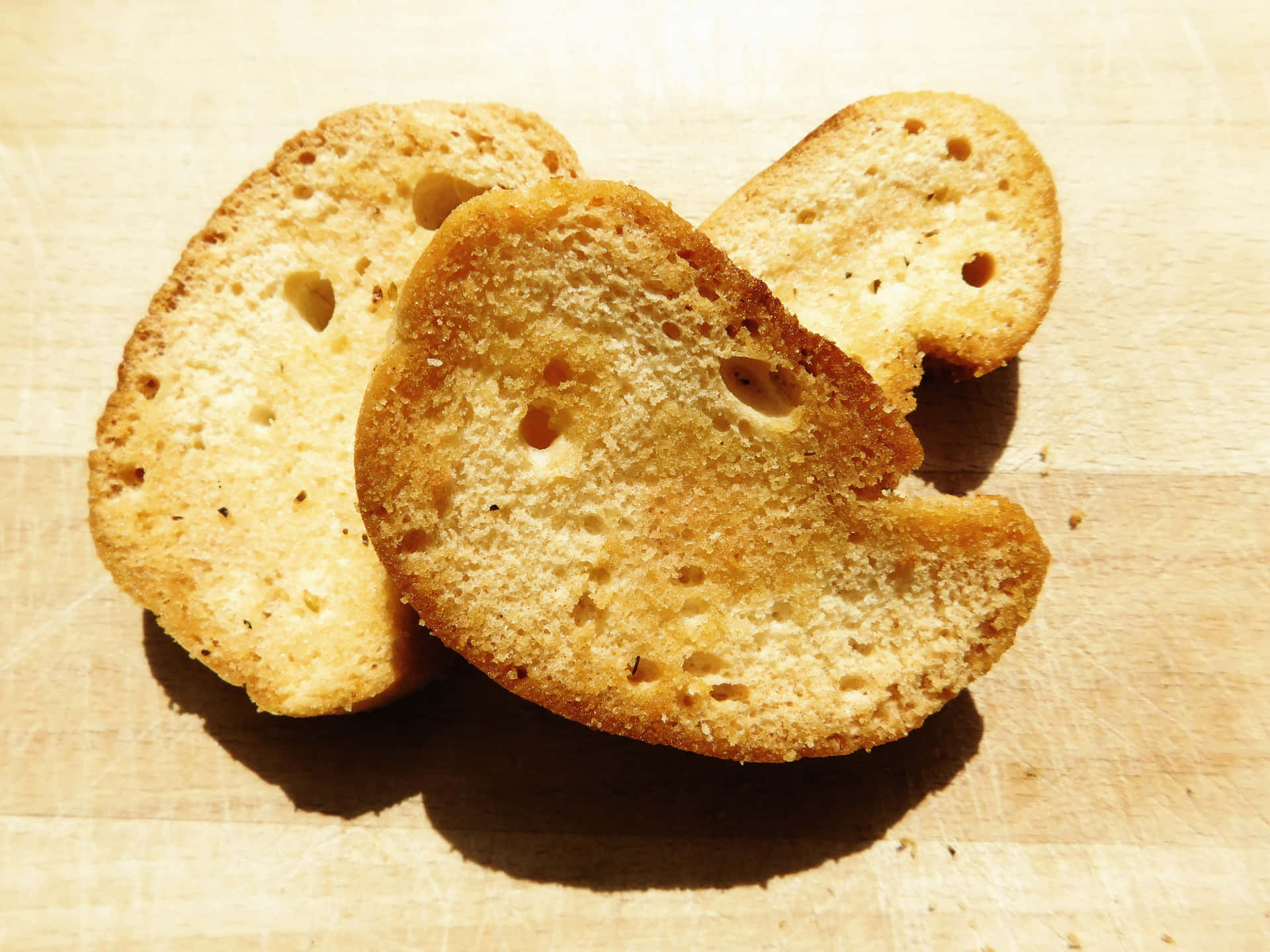 Bread 壁紙 — 三塊金黃色的烤麵包片，帶有可見的孔洞，放在淺色木質表面上，在明亮的陽光下。