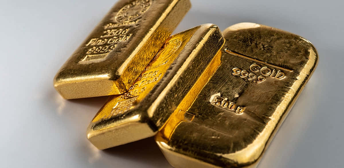 Gold Bars 桌布 壁紙 — 三塊閃閃發光的金條，帶有雕刻標記，放置在淺灰色表面上，反射出溫暖的金屬光。