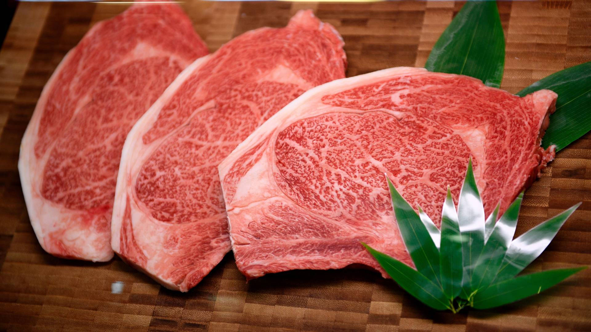 meat 桌布 壁紙 — 三塊頂級大理石紋牛排放在木板上，旁邊是新鮮的綠色竹葉。